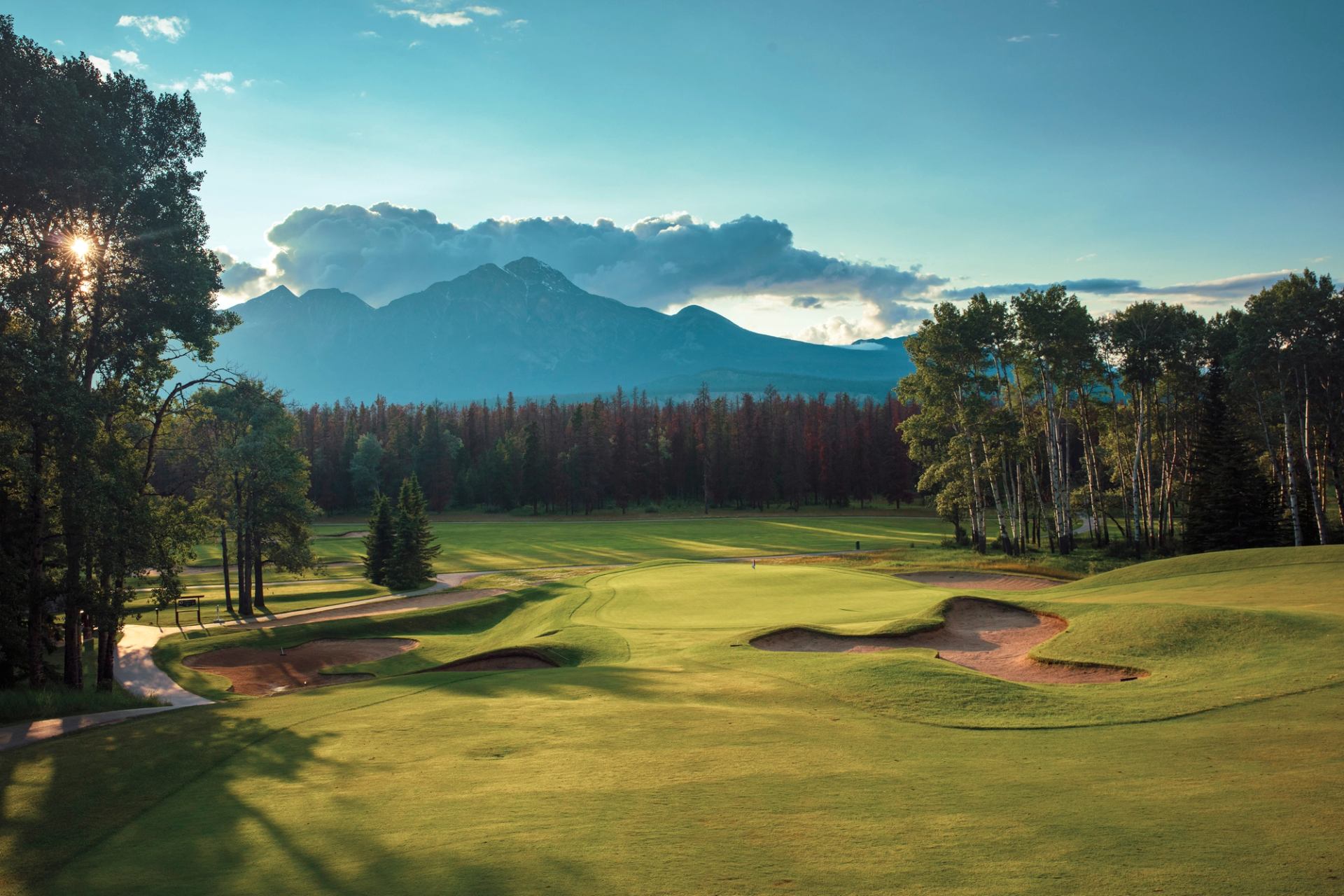 Golf course fairway with sand bunkers, surrounded by trees and distant mountains.