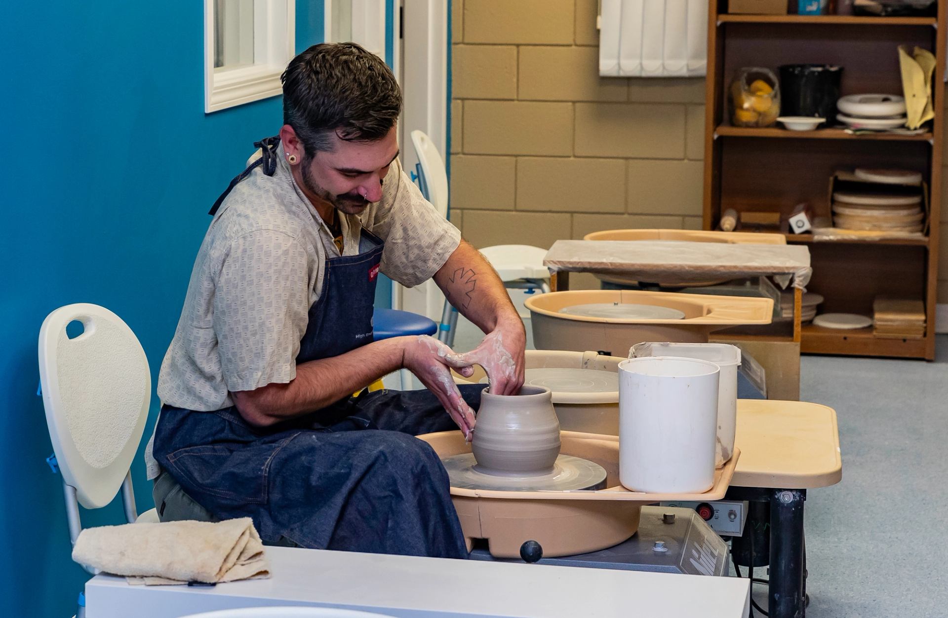 Person shaping clay on pottery wheel in studio with shelves of ceramic pieces.
