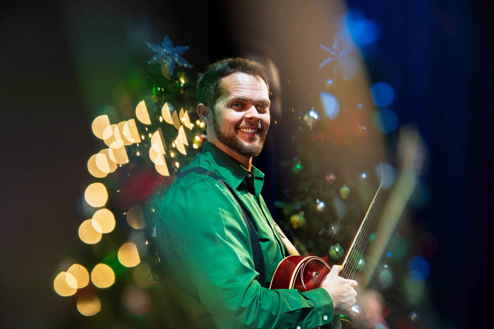 Guitarist in a green shirt performing on stage with blurred festive lights in the background.