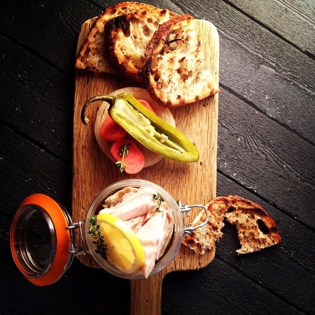 A wooden board with sliced grilled bread, pickled peppers, radishes, and a jar containing lemon slices, and thyme, set on a dark table.
