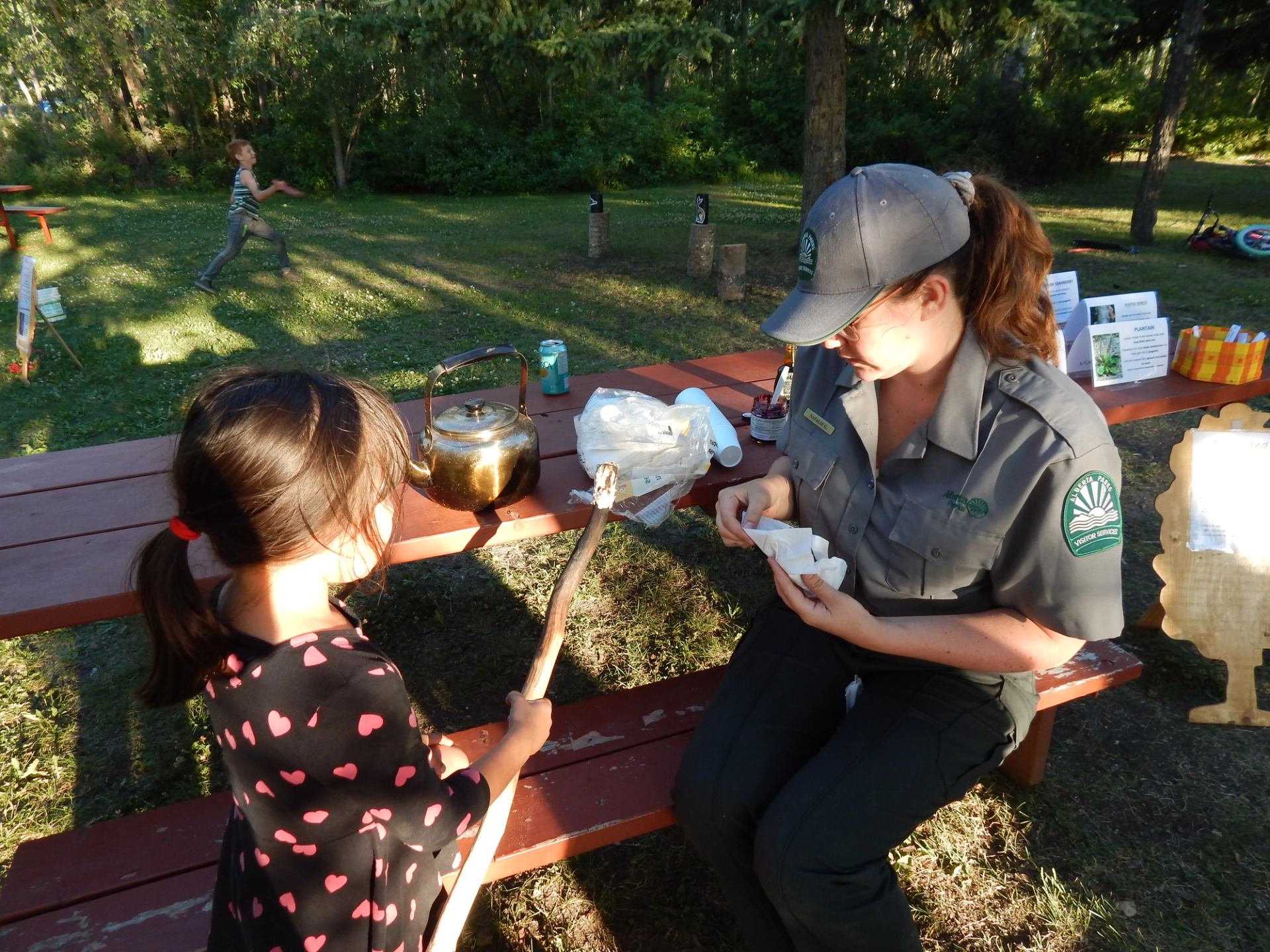 Park staff member helps child roast marshmallow at picnic table in grassy campground.