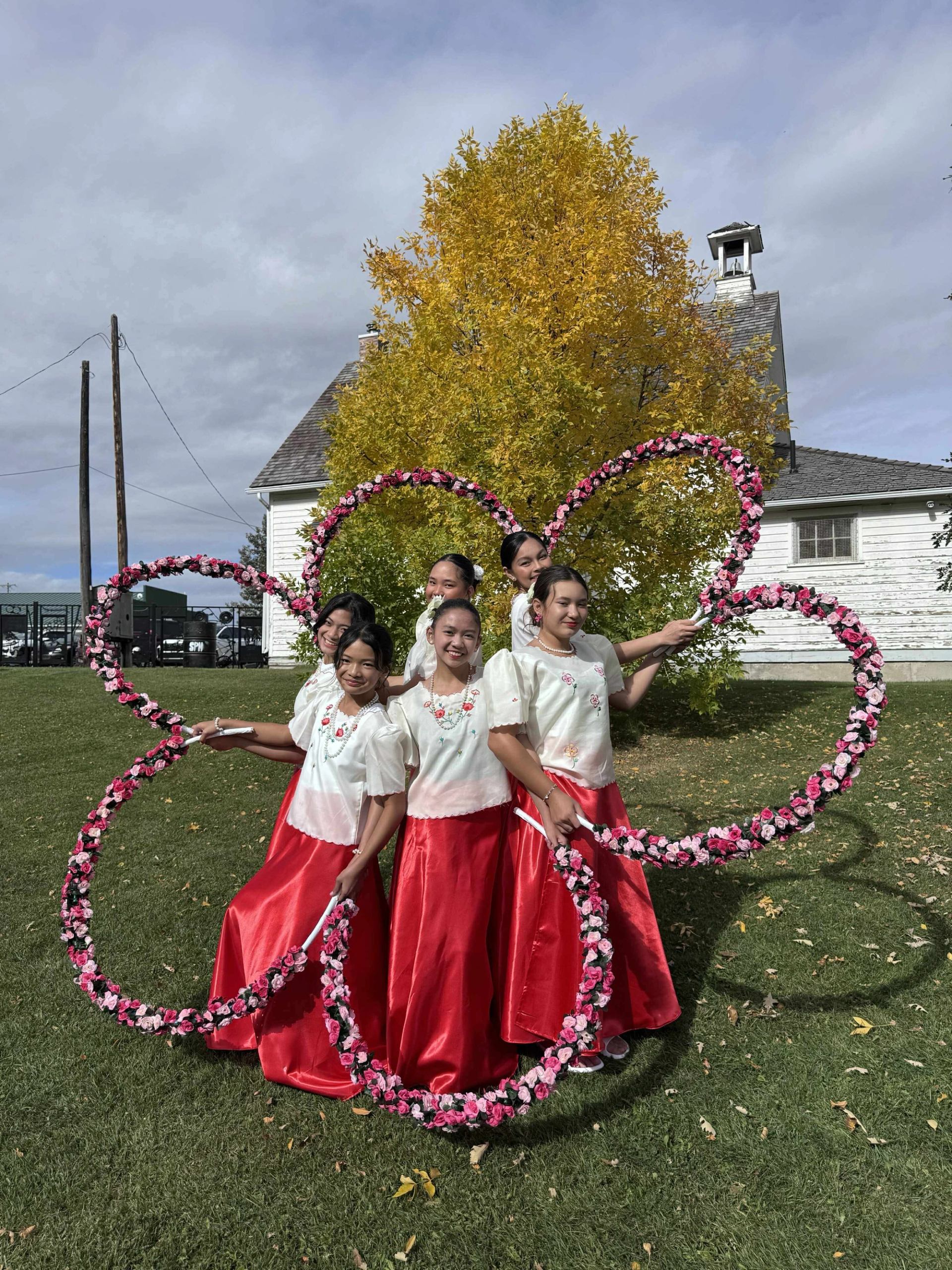 Dancers in red skirts hold pink floral hoops on grass before yellow tree and building.