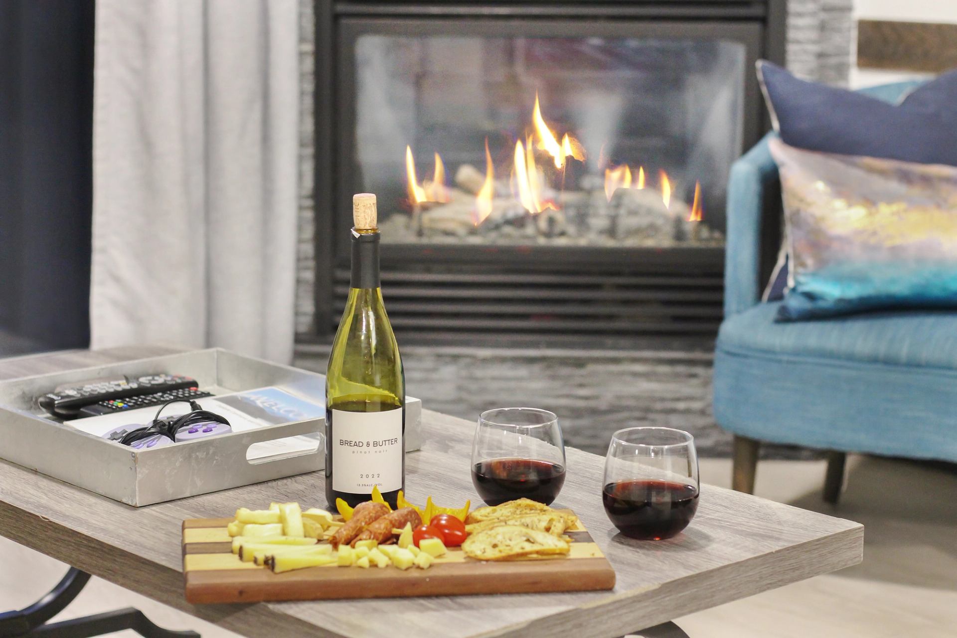 Wine, two glasses and a charcuterie board sit before a lit fireplace.