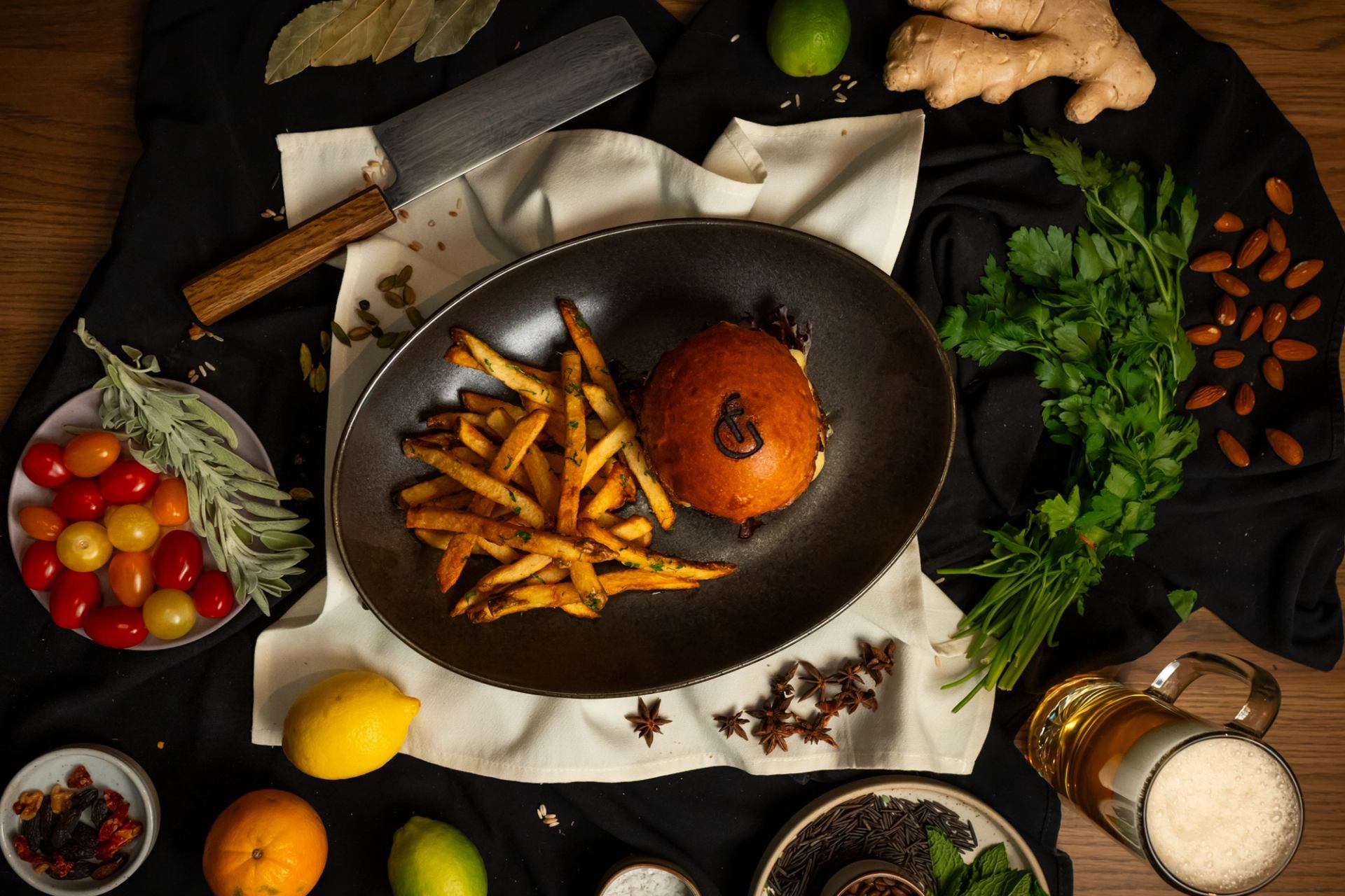 Burger with Good Folk logomark burned into the bun with house cut fries on a black plate arranged among herbs an vegetables