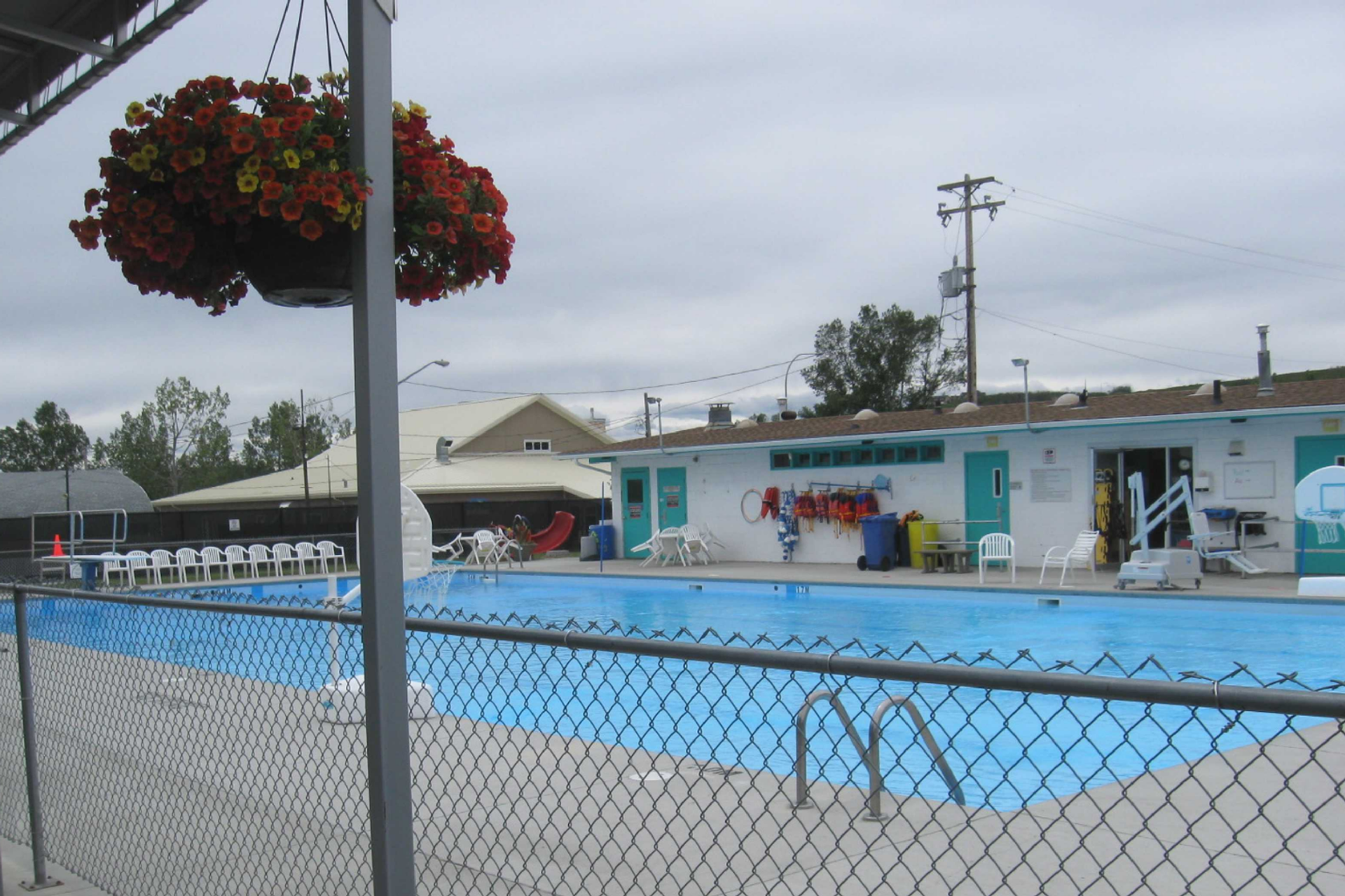Diamond Valley Municipal Campground features a clean outdoor swimming pool enclosed by a fence, with white chairs, a building with amenities, and colorful hanging flower baskets nearby. The scene presents a well-maintained and welcoming space for relaxation and swimming.