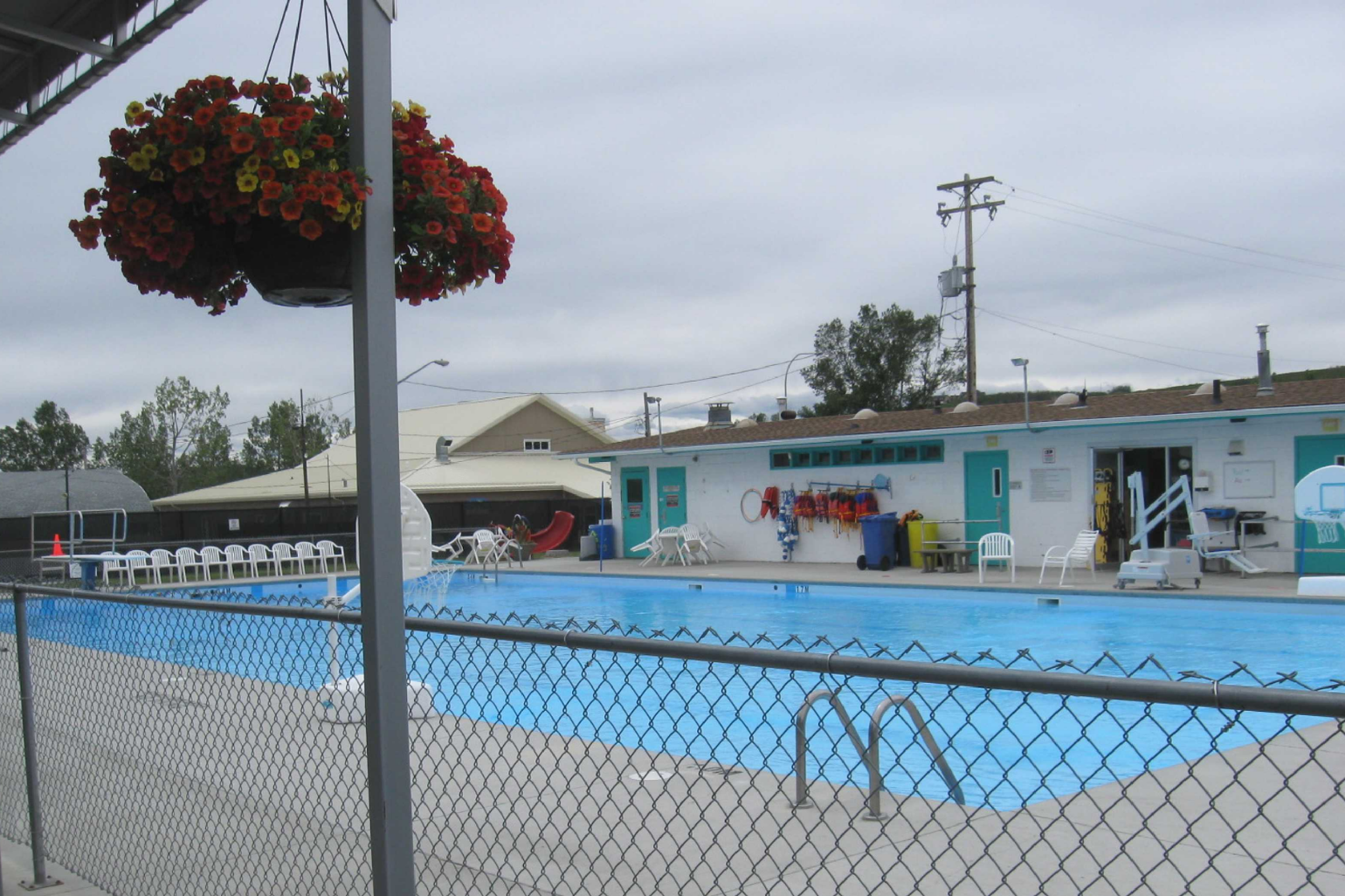 Diamond Valley Municipal Campground features a clean outdoor swimming pool enclosed by a fence, with white chairs, a building with amenities, and colorful hanging flower baskets nearby. The scene presents a well-maintained and welcoming space for relaxation and swimming.