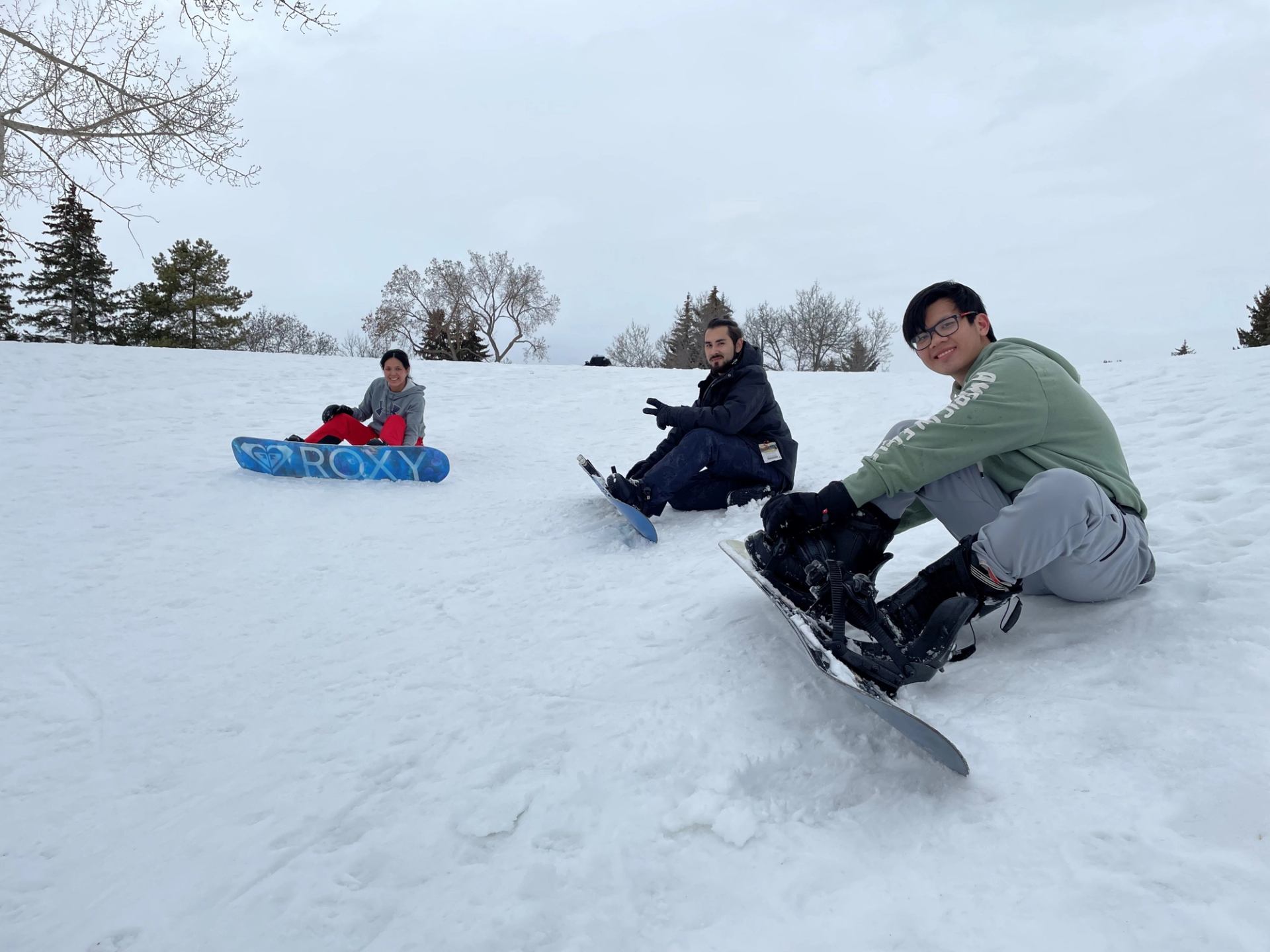Snowboarders crouch on snow in a park, holding boards and posing for camera.