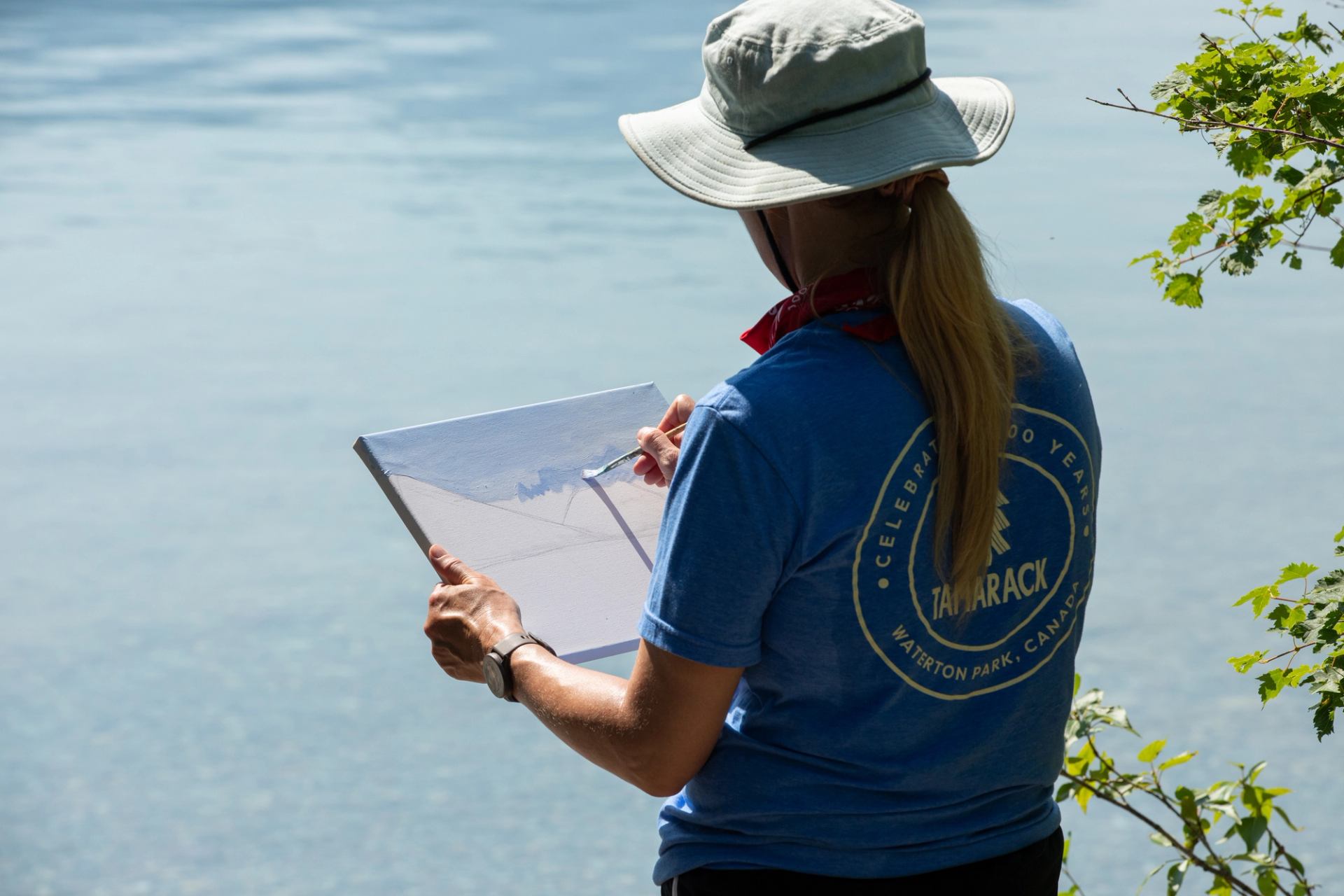 Person in Tamarack T-shirt paints on canvas beside calm lake, wearing sun hat.