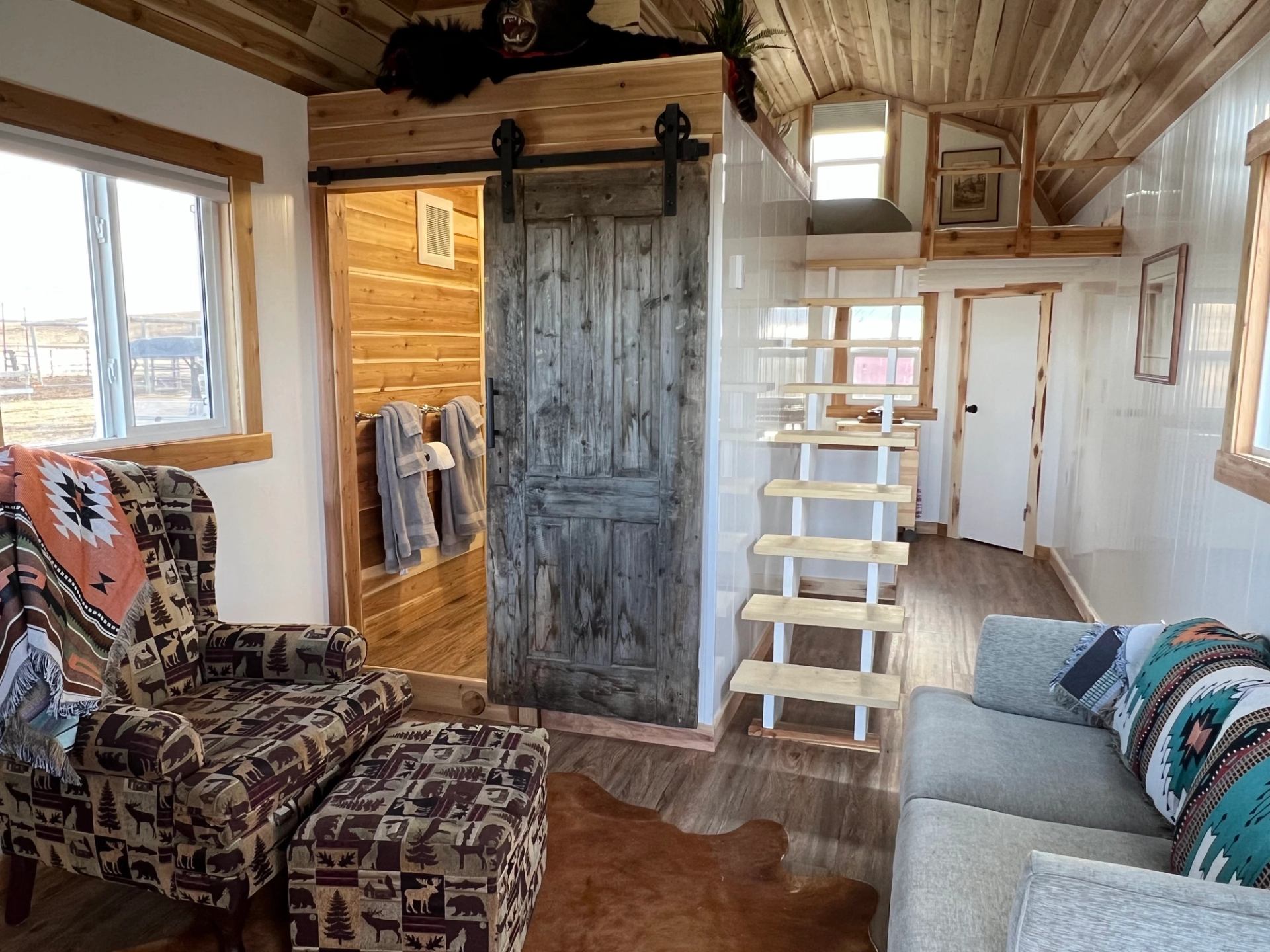Cabin interior with wood walls, patterned chairs, sofa, and a loft accessed by wooden steps.