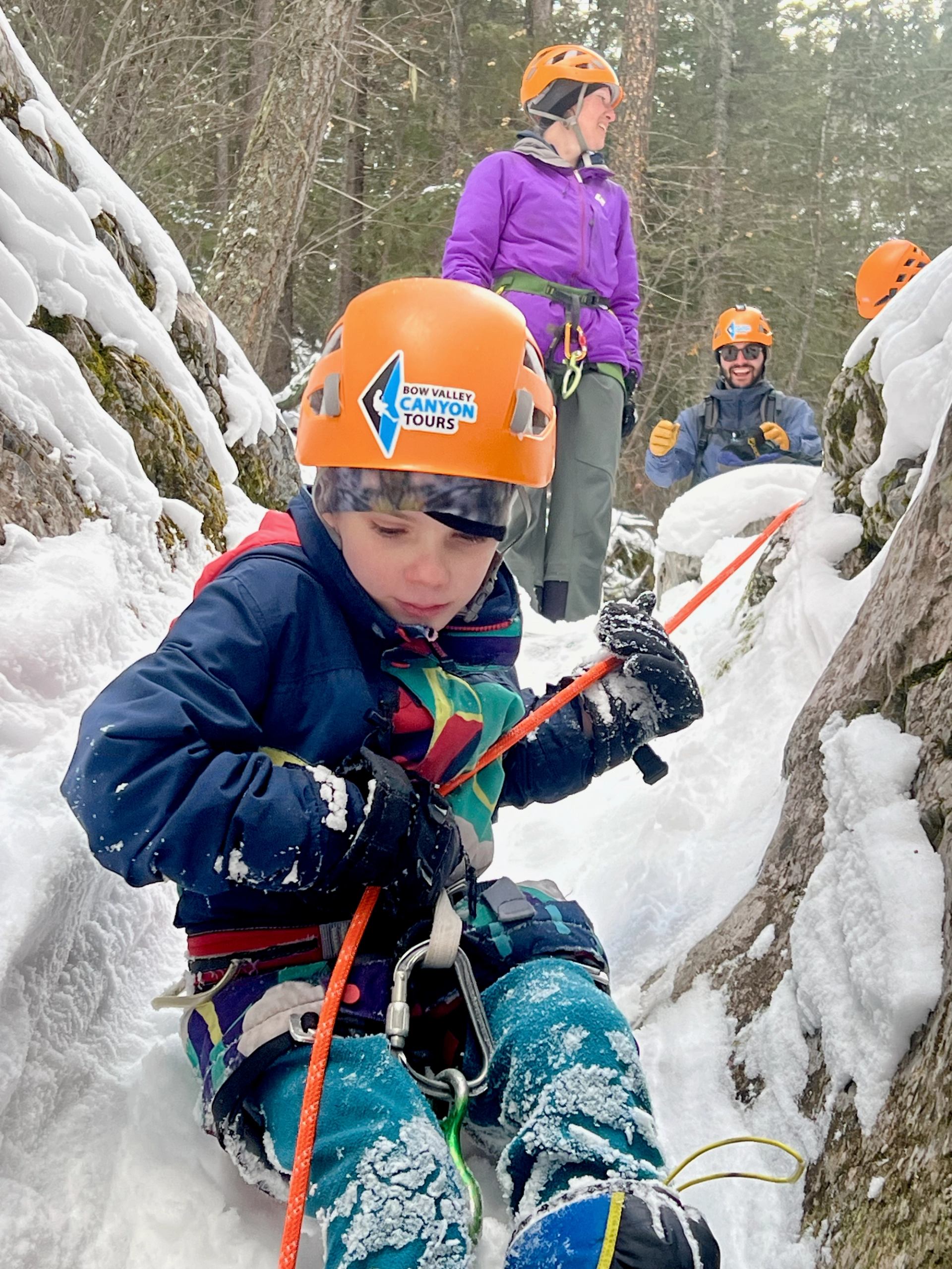 Canyon tour participant in orange helmet, gloved hands on rope in snowy canyon rocks.