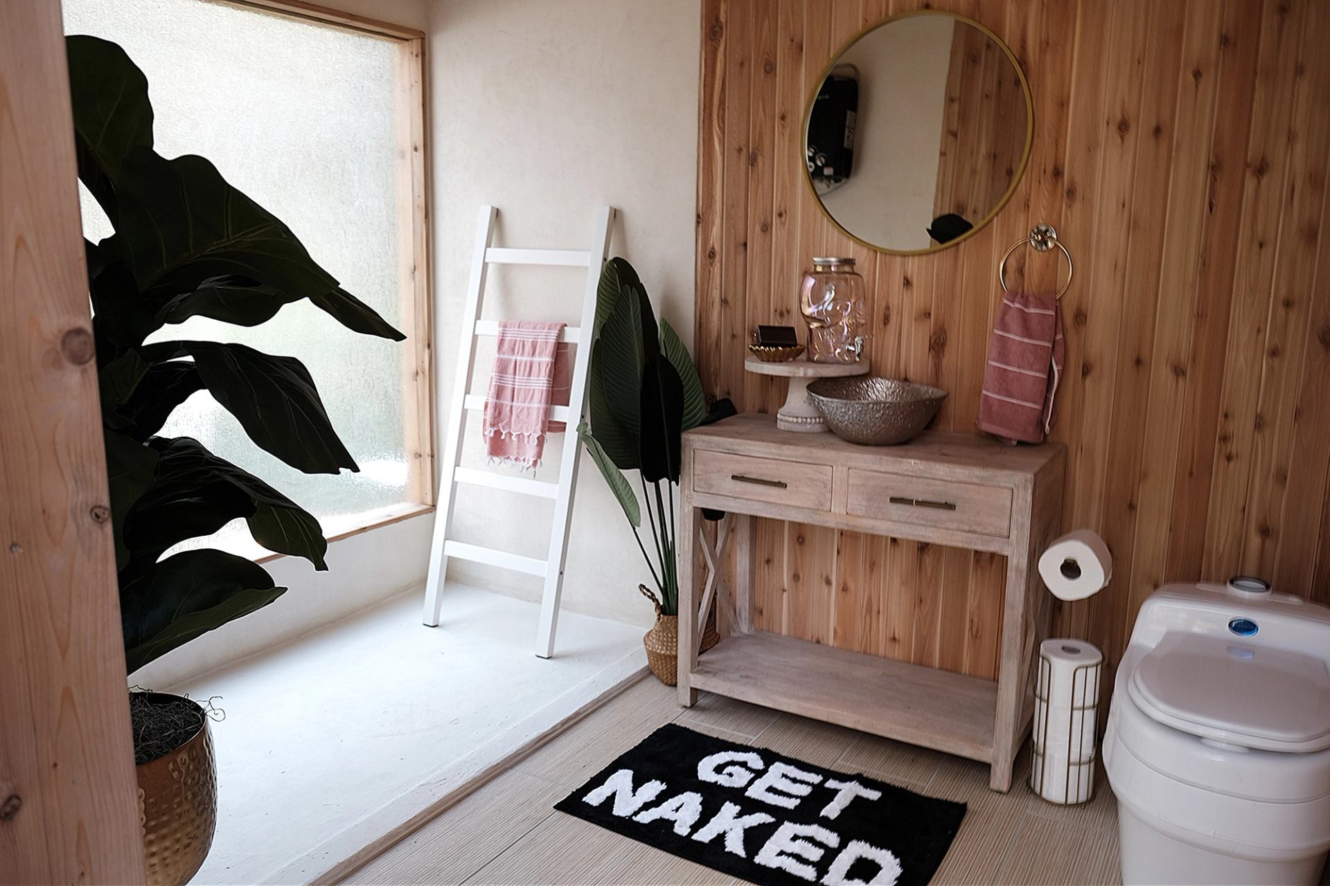 A modern bathroom with a wood-paneled wall, light wood vanity, round mirror, and large green plants.
