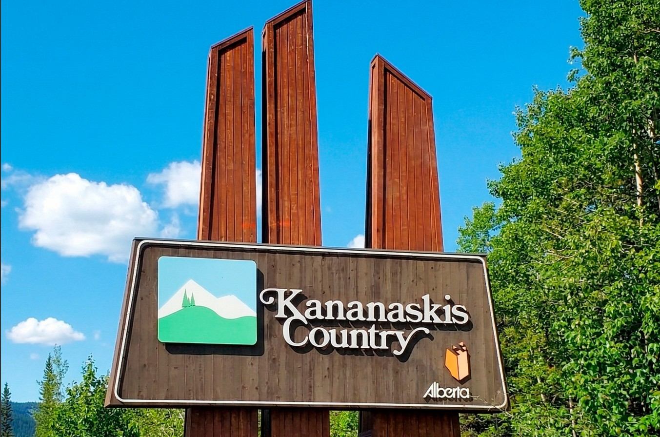 Kananaskis Country entrance sign with wood panels, Alberta logo, and green trees under blue sky.