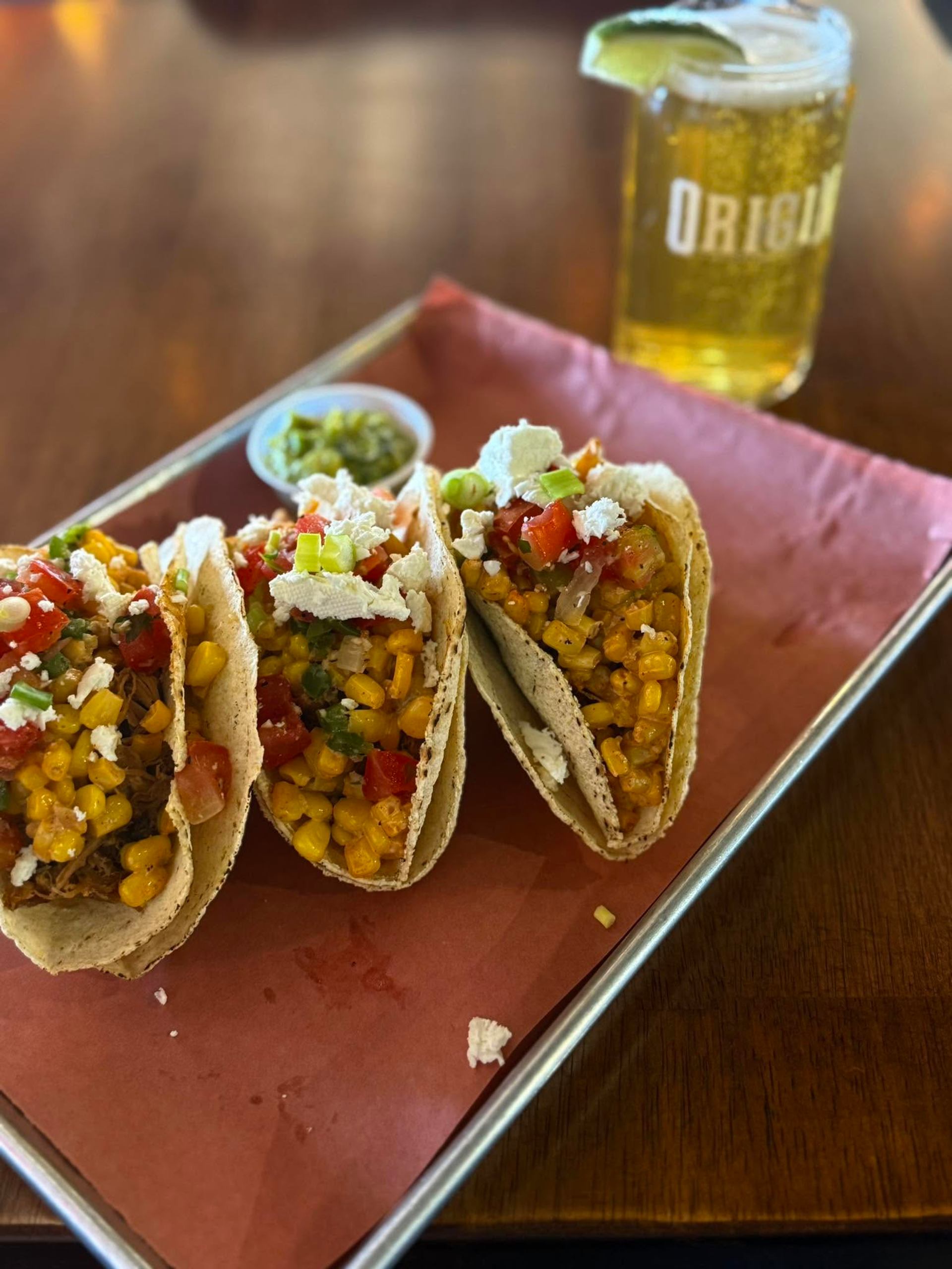 Three tacos filled with corn, vegetables, and cheese are served beside a cold beverage.