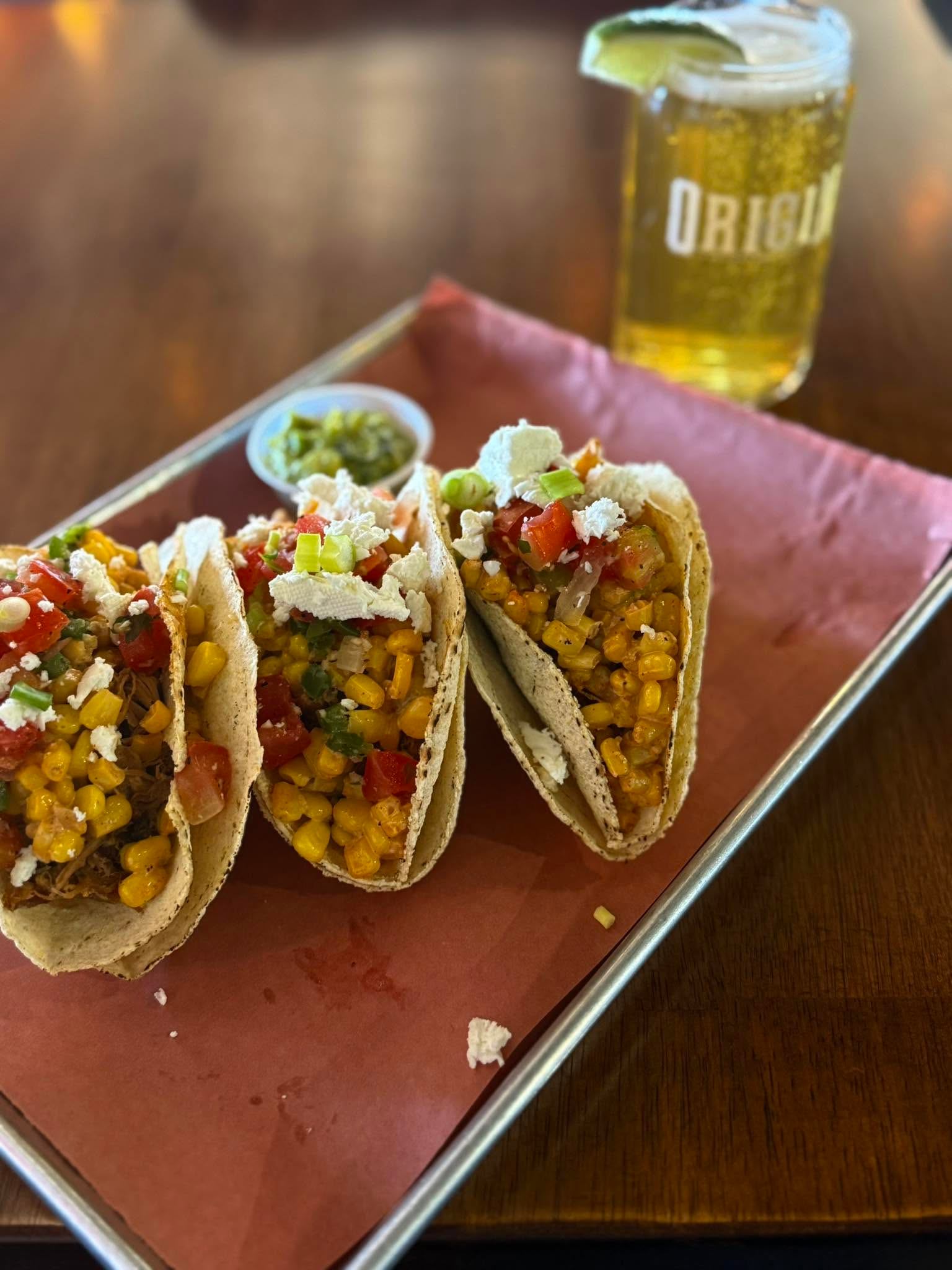 Three tacos filled with corn, vegetables, and cheese are served beside a cold beverage.