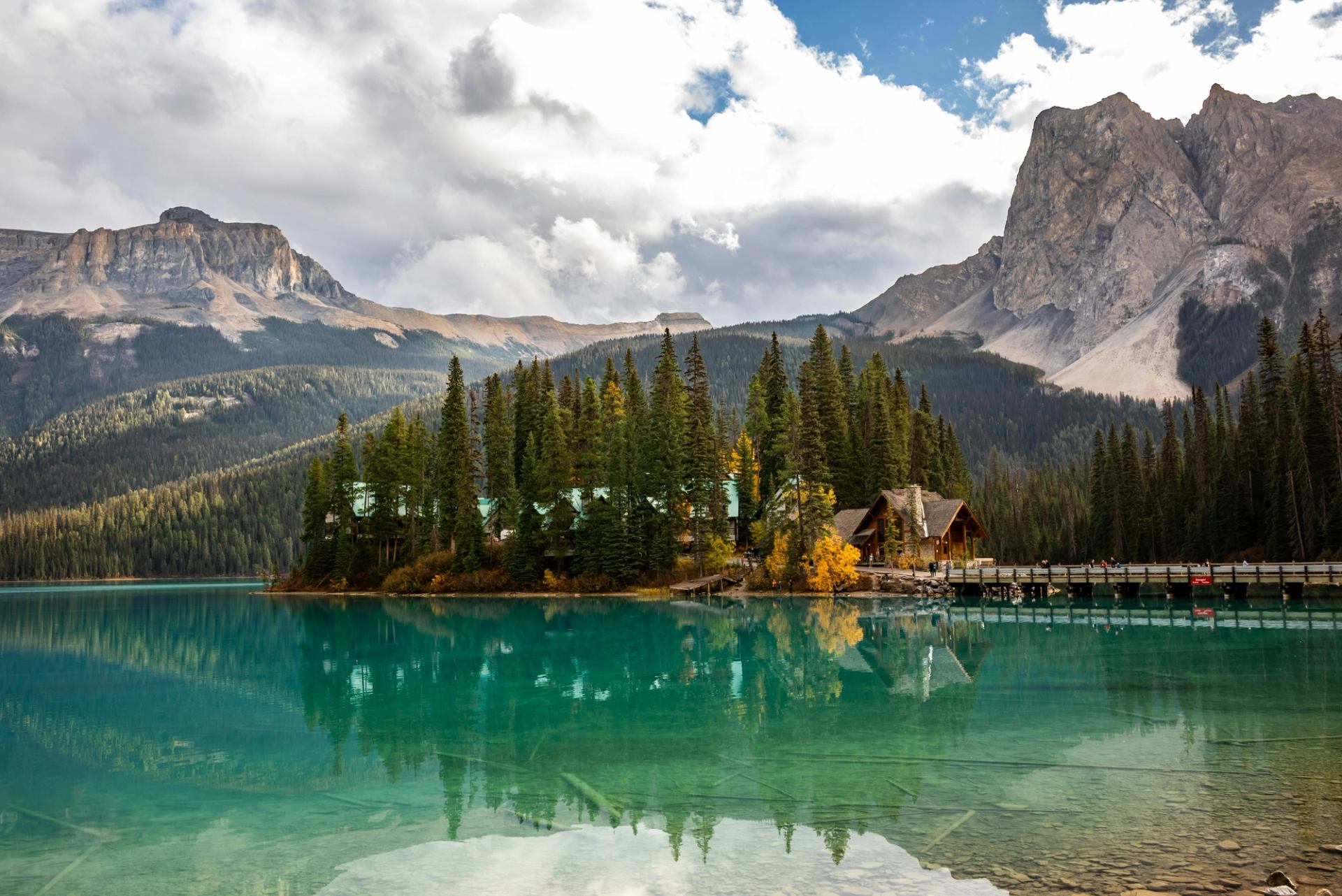 Turquoise lake reflects forested island, wooden lodge, bridge and surrounding rocky mountains.