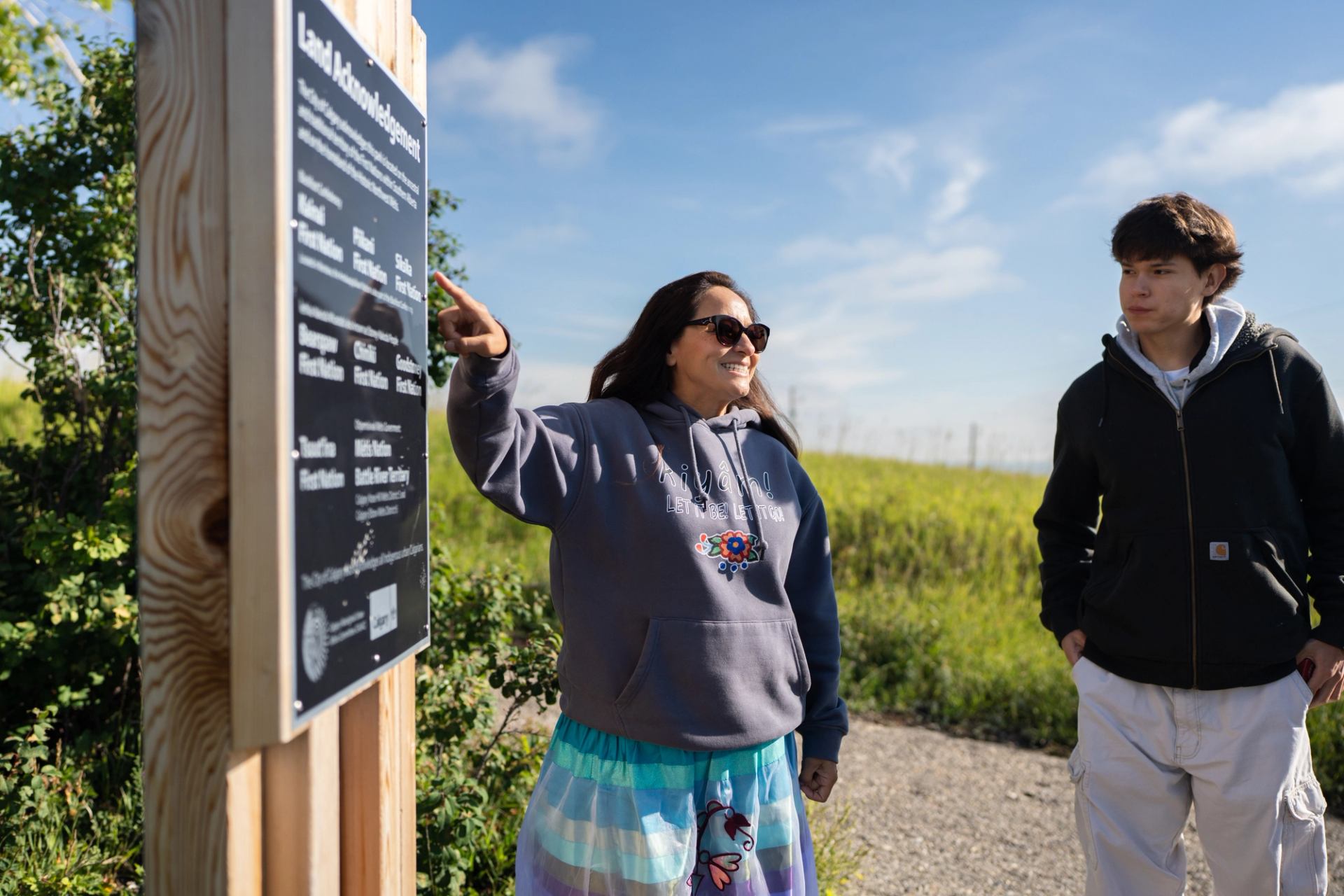 Two people stand near an outdoor sign along a sunny gravel path.