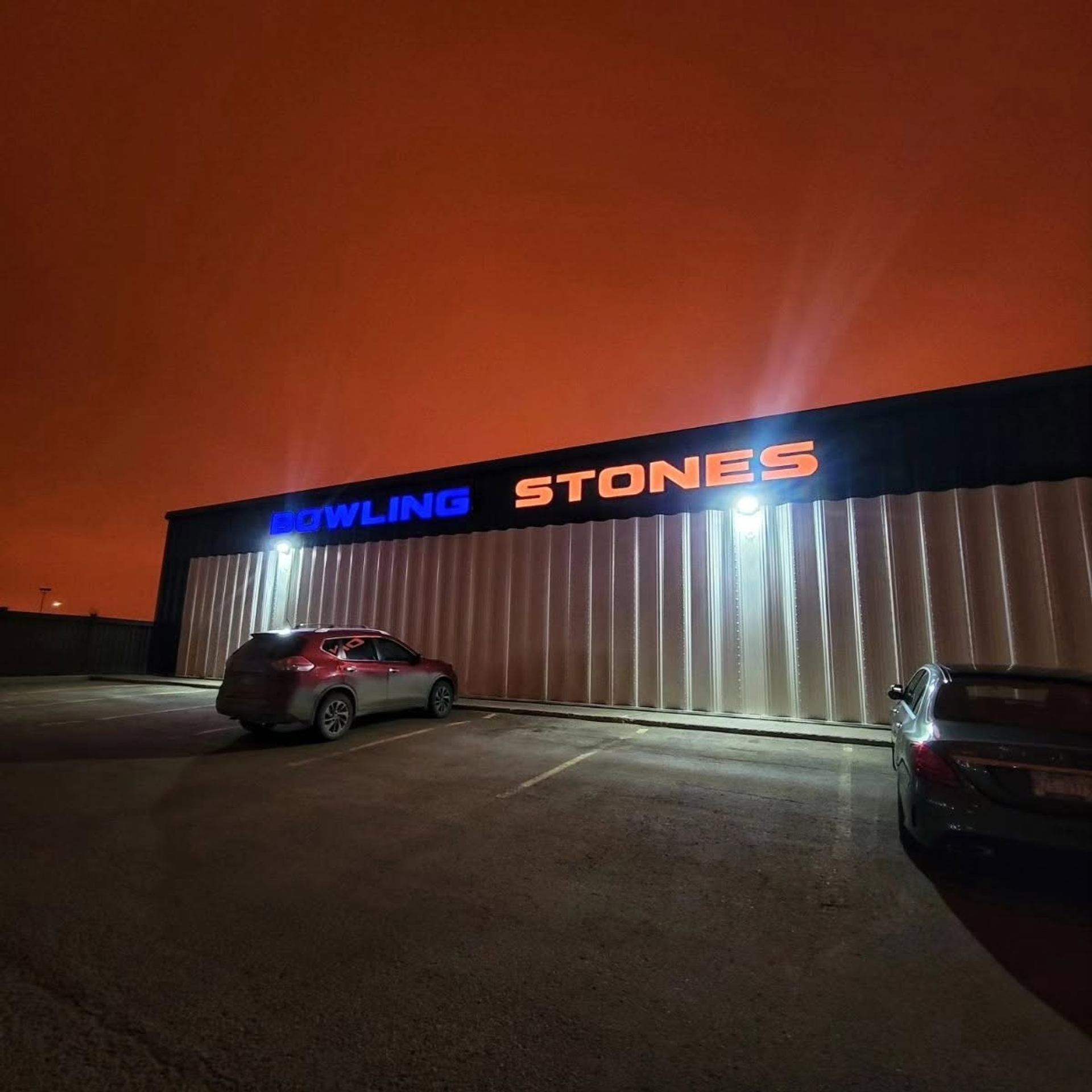Parking lot outside Bowling Stones building under orange sky, with two parked cars.