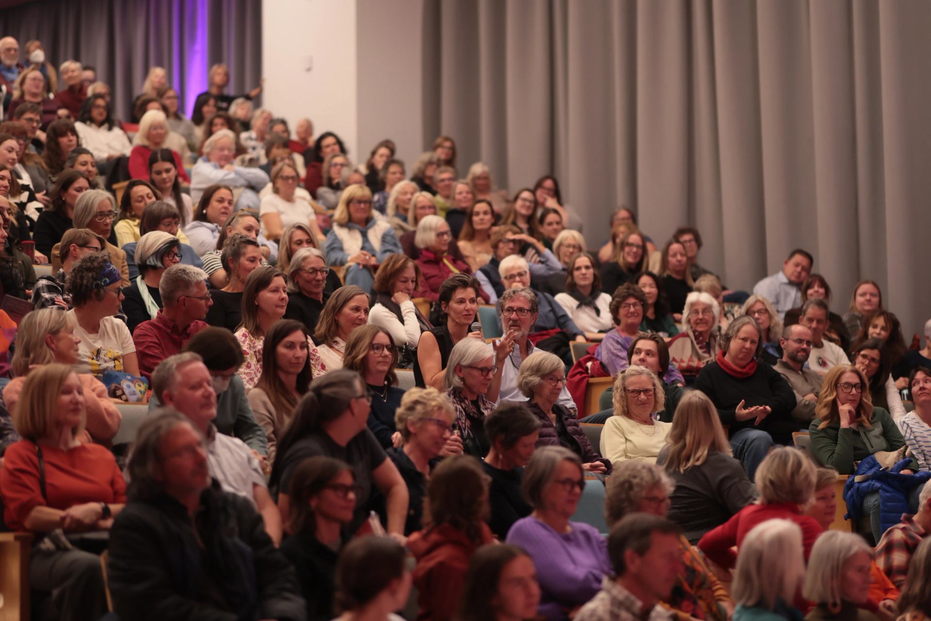 Audience members sit in tiered theatre seating beside grey curtains.
