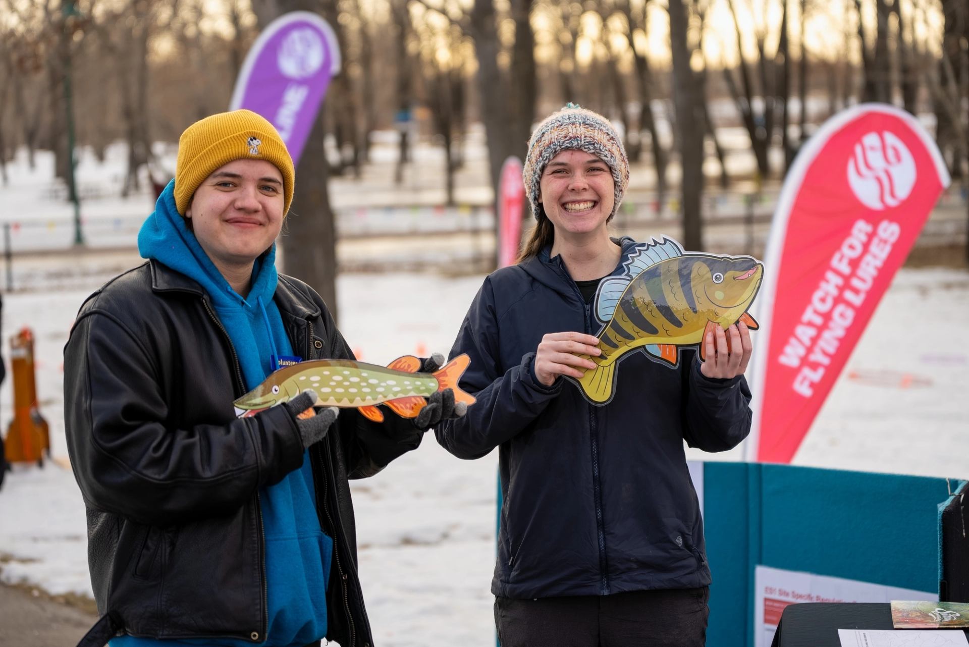 Two people holding fish-shaped cutouts at an outdoor Fishtival event with banners in the background.