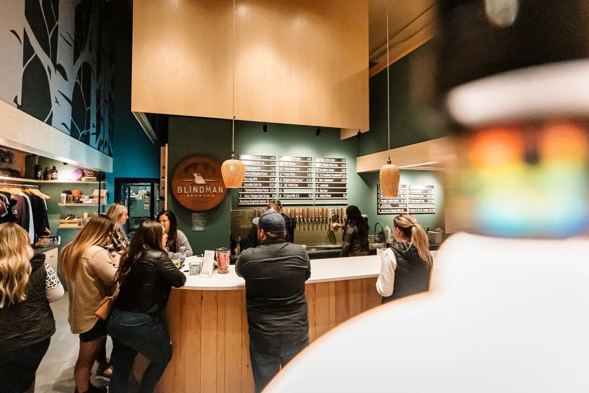 Visitors gather at a taproom bar with beer taps and a menu board in the background.