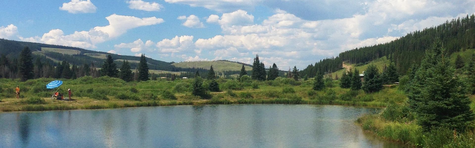 People fishing under umbrella near pond, with forested hills and blue sky in the background