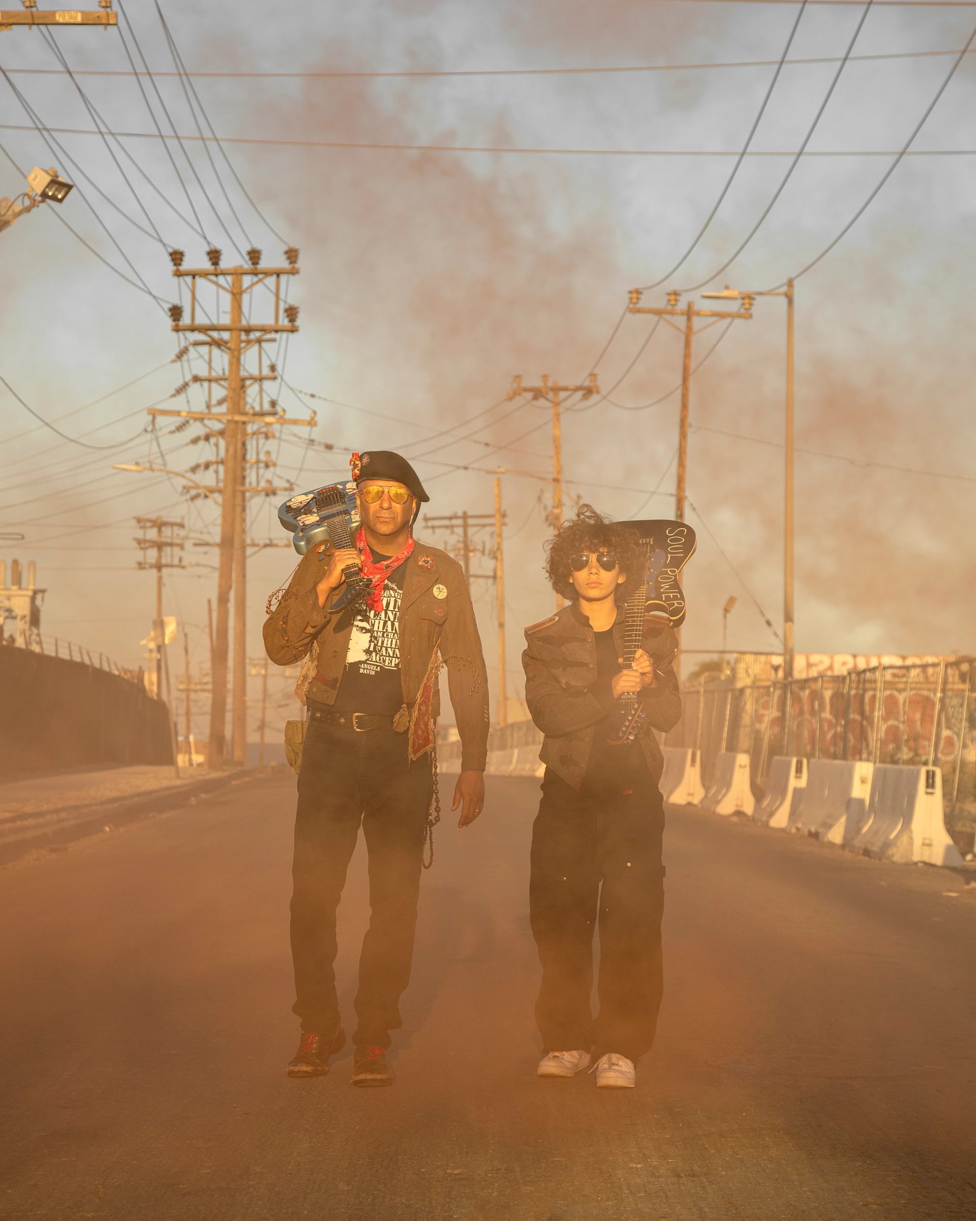 Two people walk down a dusty street carrying electric guitars amid power lines.