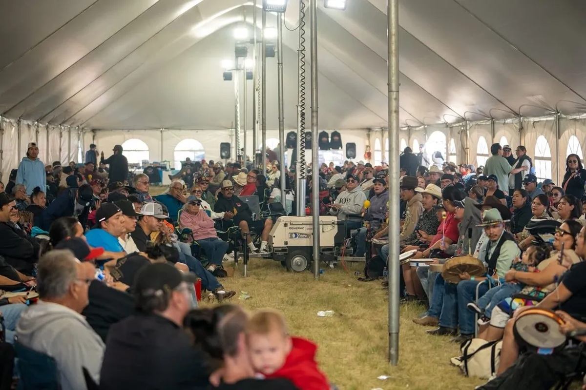 Large crowd sits under a white event tent, listening and drumming during gathering.
