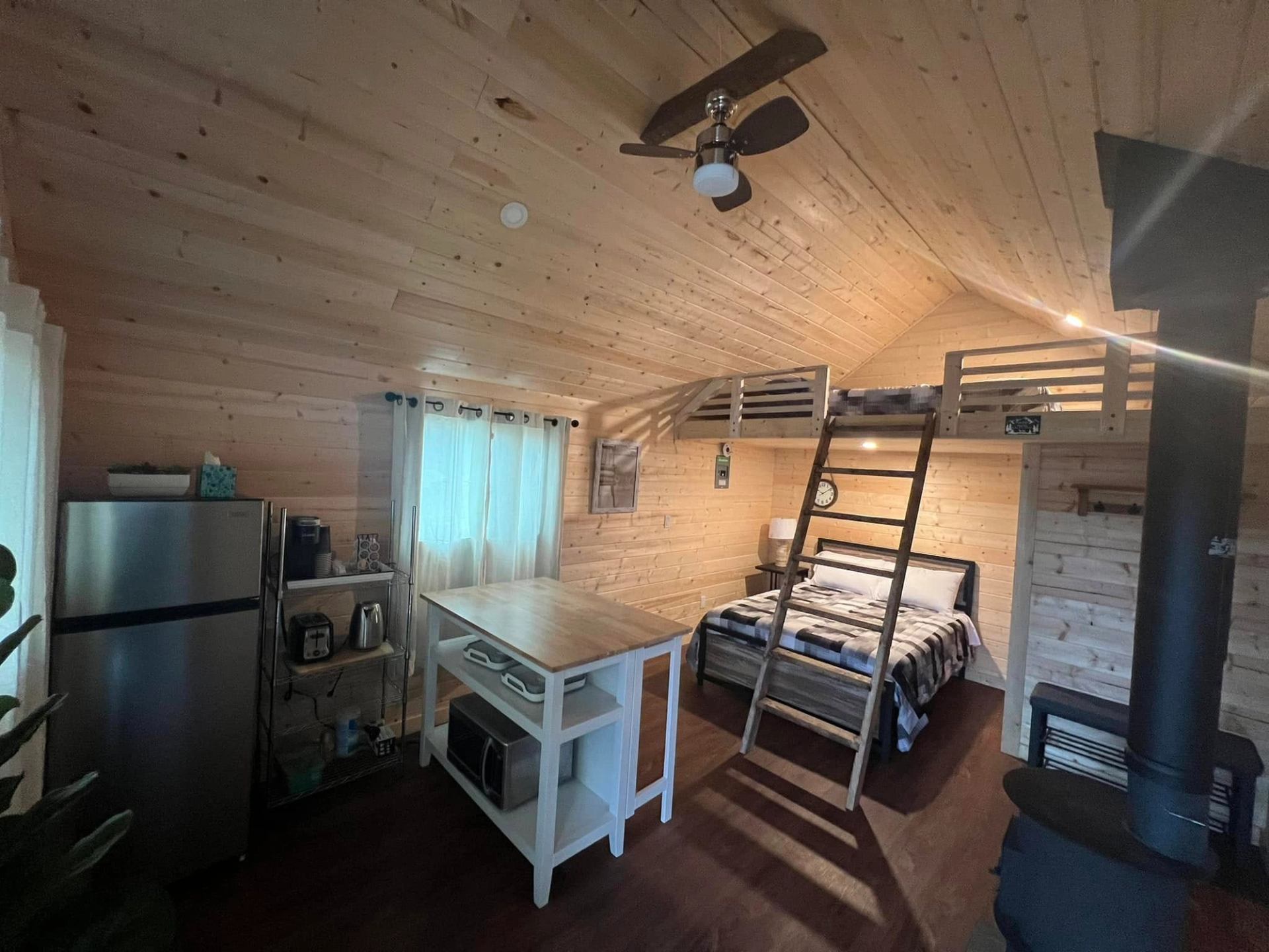 Wood-panelled tiny cabin interior with loft bed, kitchen area, island and wood stove.