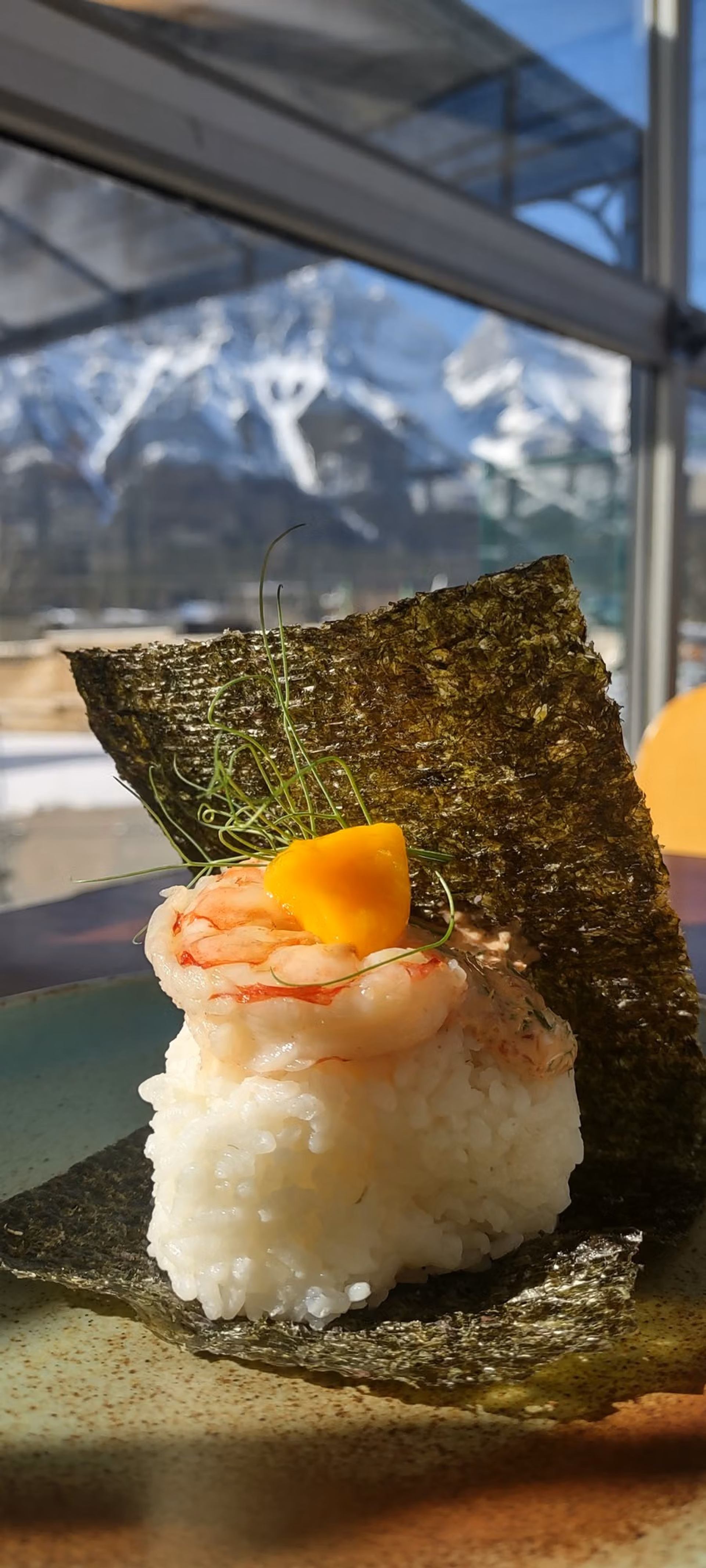 A Japanese rice-based delicacy topped with shrimp and garnish is served beside crisp seaweed and mountain views.