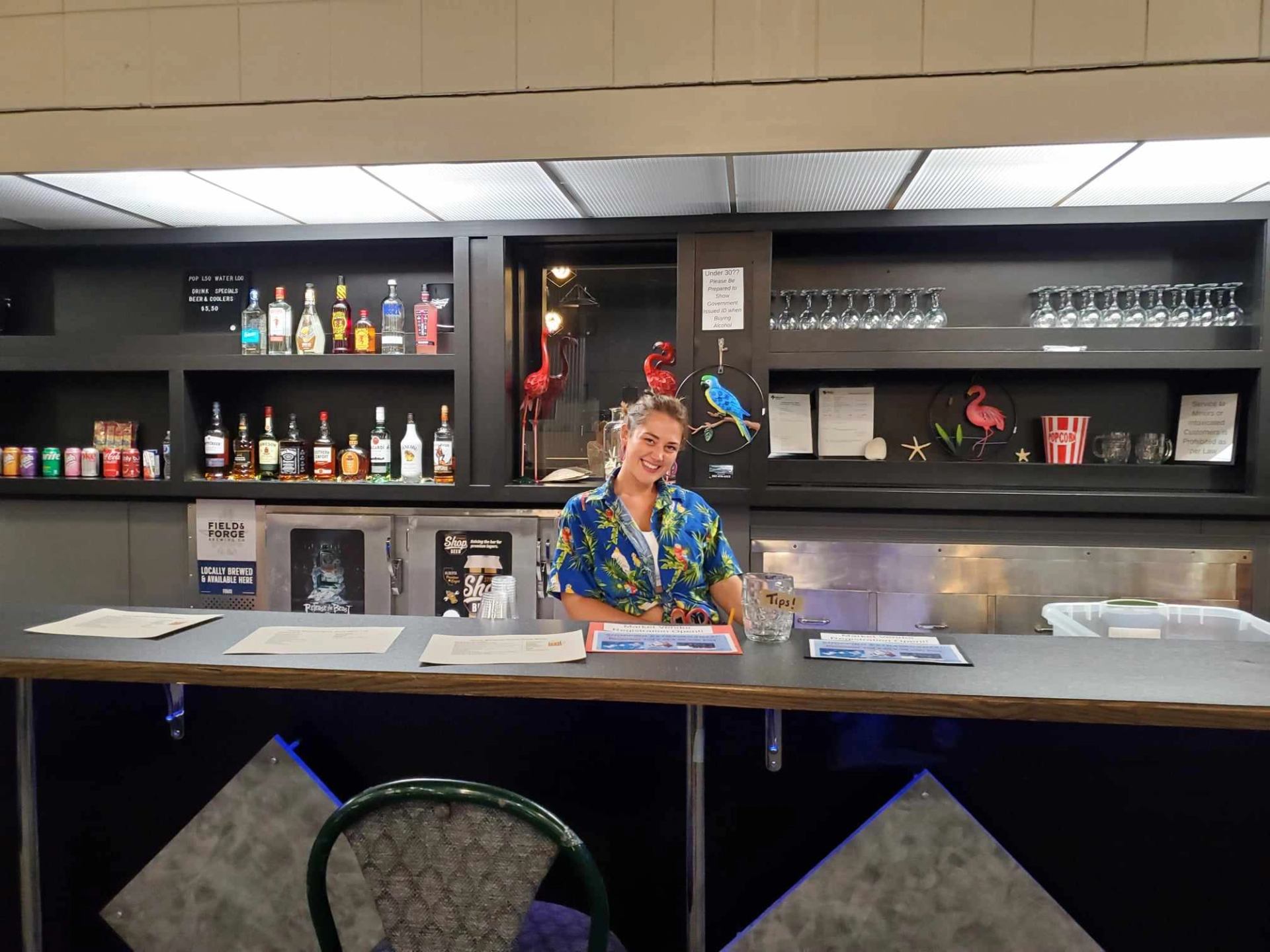 Bartender in floral shirt stands behind bar with liquor bottles, glasses and tip jar.