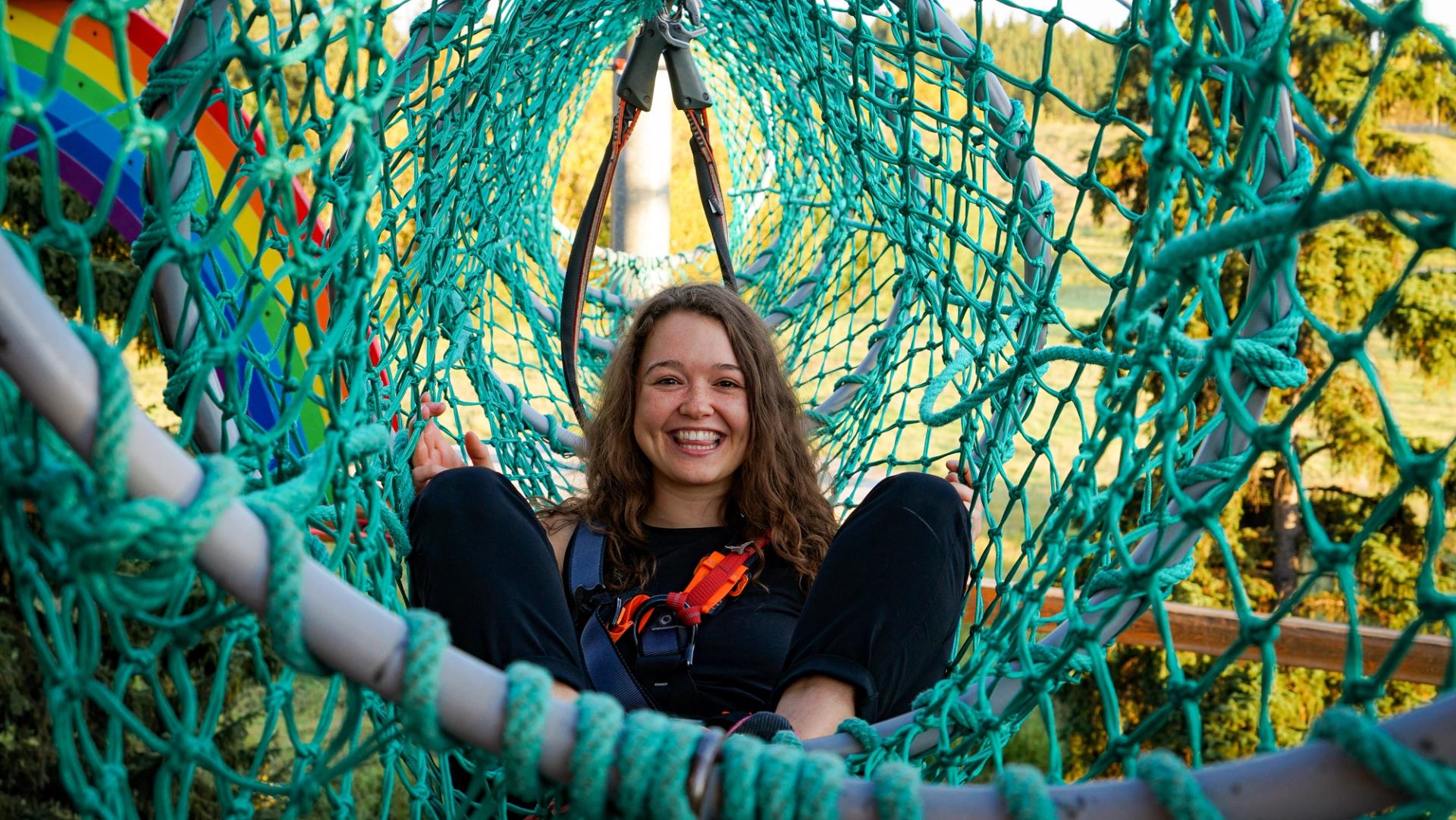 A woman smiles and relaxes on a net-style climbing tower obstacle.
