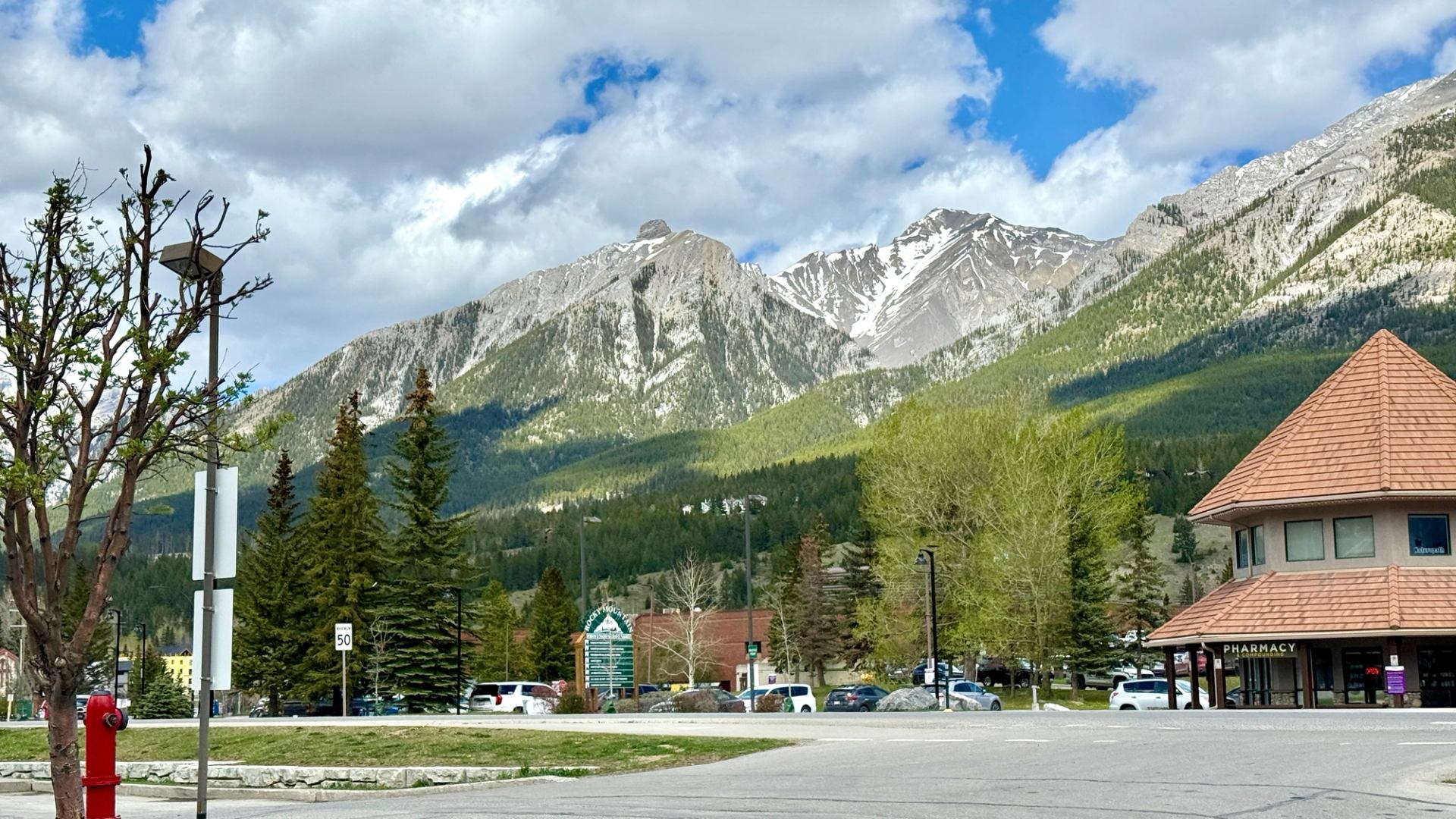 Mountain peaks rise behind trees, parked cars and a pharmacy building.