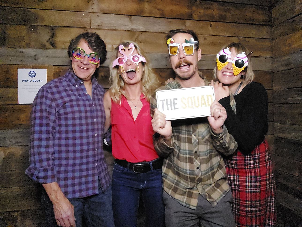 Four people in novelty glasses pose in front of a wooden wall, one holding a “THE SQUAD” sign.