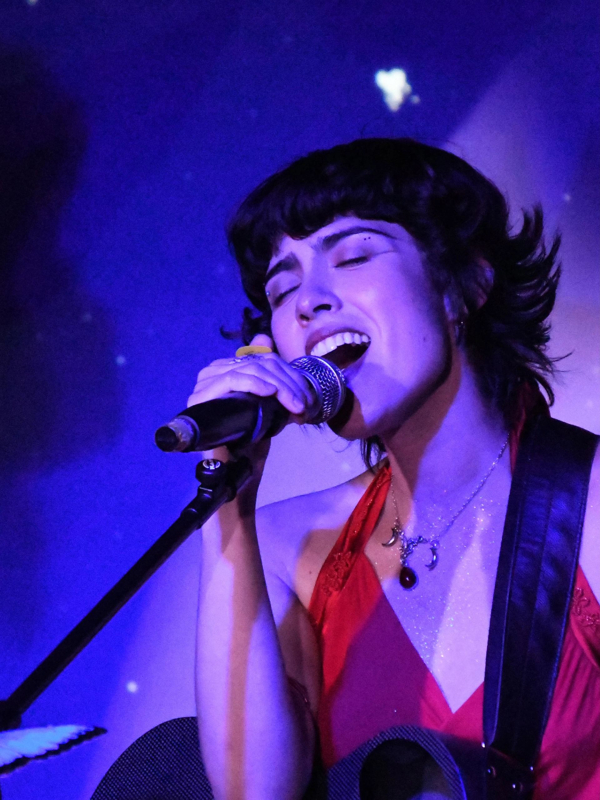 Singer in a red top performs with a microphone under purple stage lighting.
