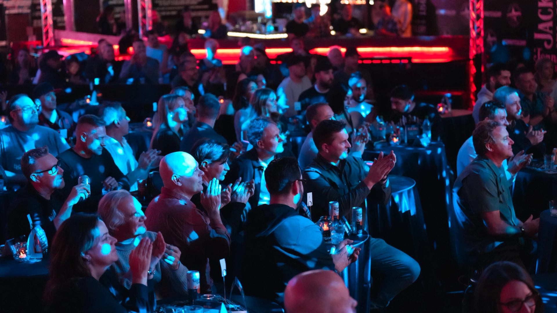 A seated audience claps and watches a comedy performance in a dimly lit venue with blue and red lighting.