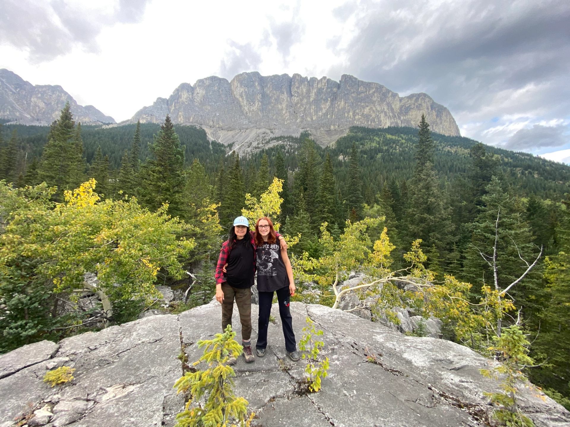 Two hikers stand on a rocky overlook surrounded by forest with towering mountain cliffs behind them.