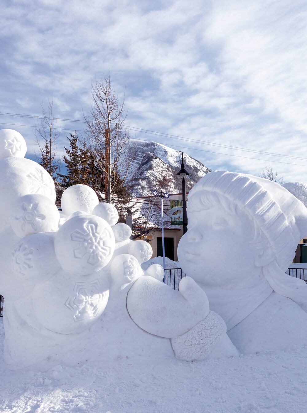 Snow sculpture of child with snowflakes, set against trees and a mountain backdrop.
