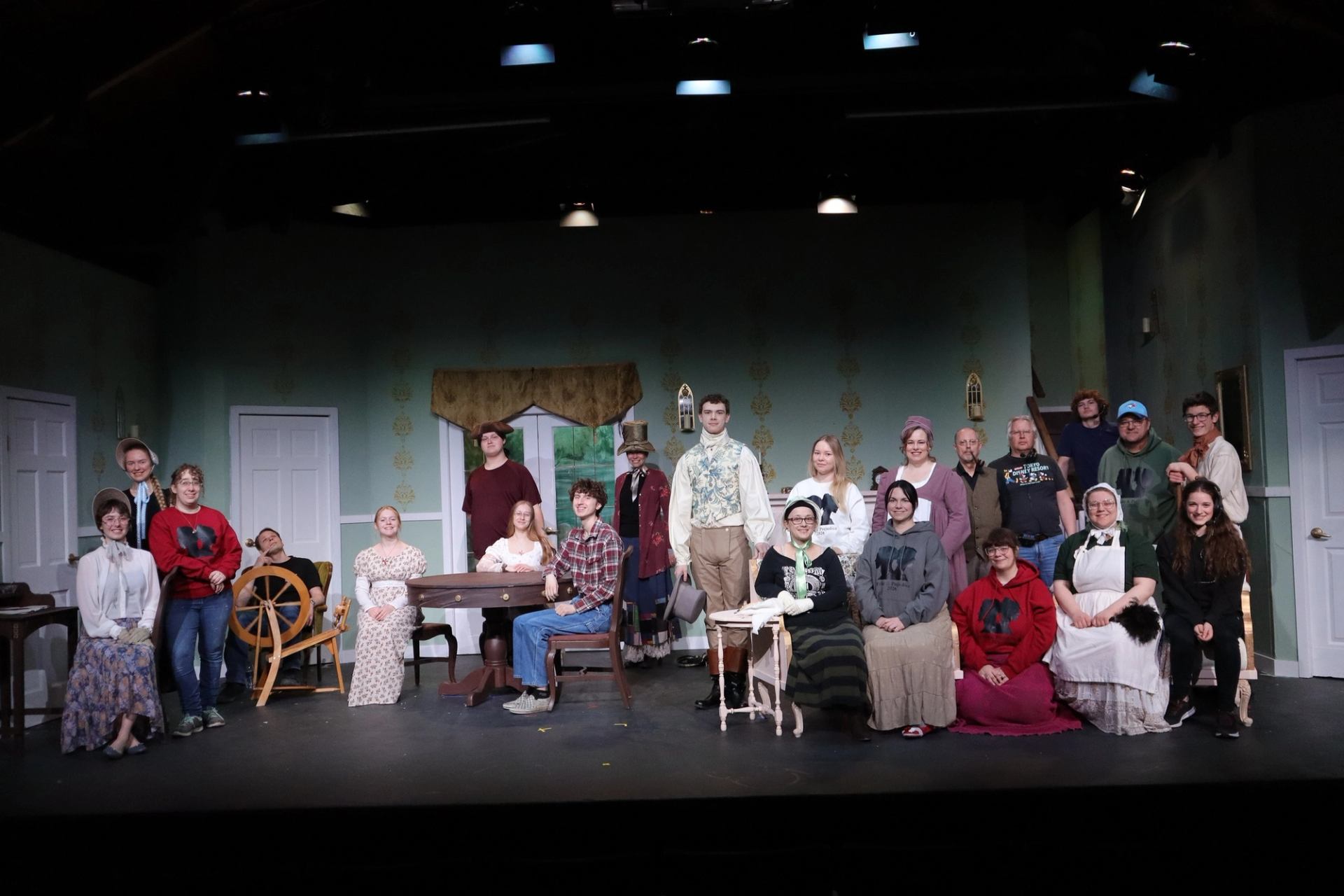 Large theatre cast poses on stage in period costumes and casual clothes.