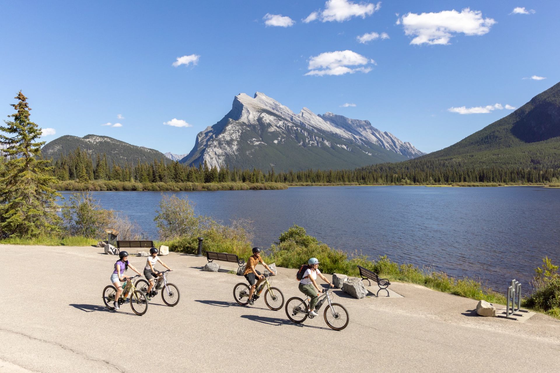 Cyclists ride along a lakeside path with forest and rocky mountain backdrop.