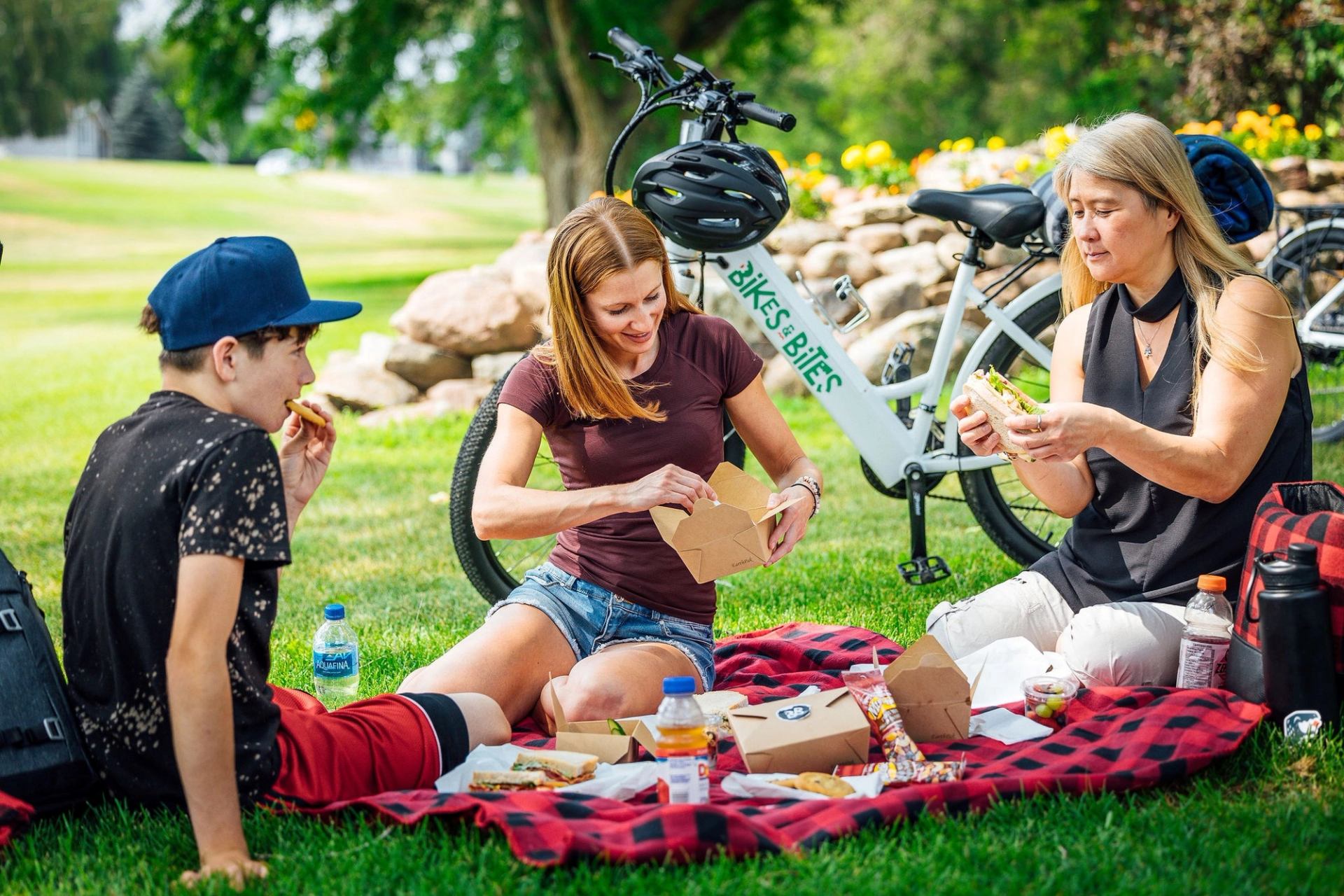 Three people enjoy a picnic on a red blanket in a park with food containers and bikes behind them.