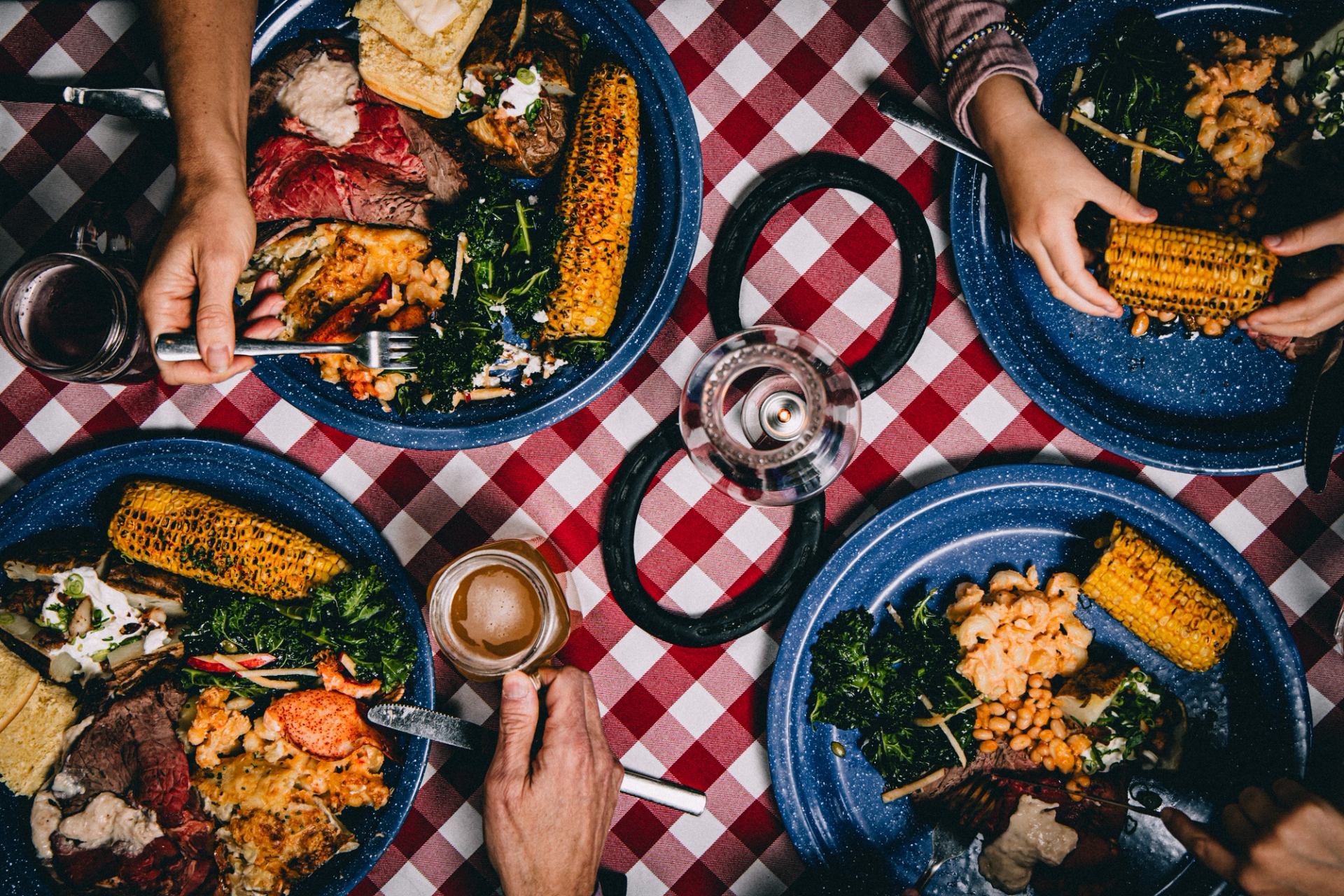 Overhead view of hands reaching for plates of roast beef, corn, and sides on a red checkered tablecloth.