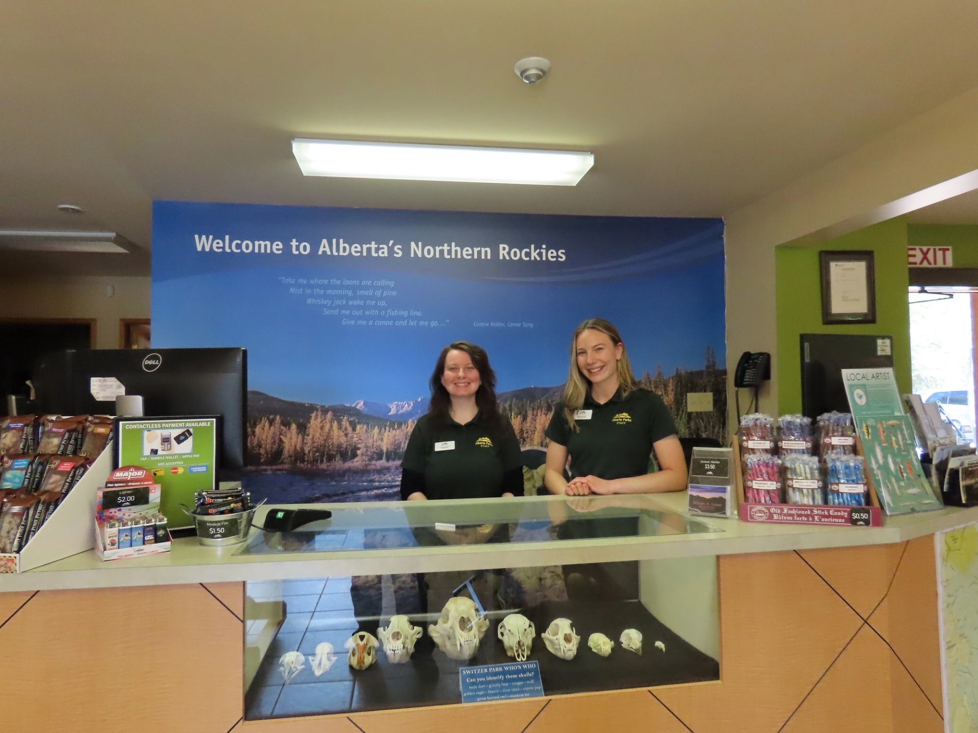 Visitor centre reception desk with two staff, brochures, snacks and Alberta’s Northern Rockies sign.
