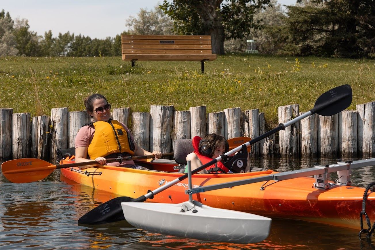 Two people kayaking together on calm water near a grassy shore and wooden barrier.