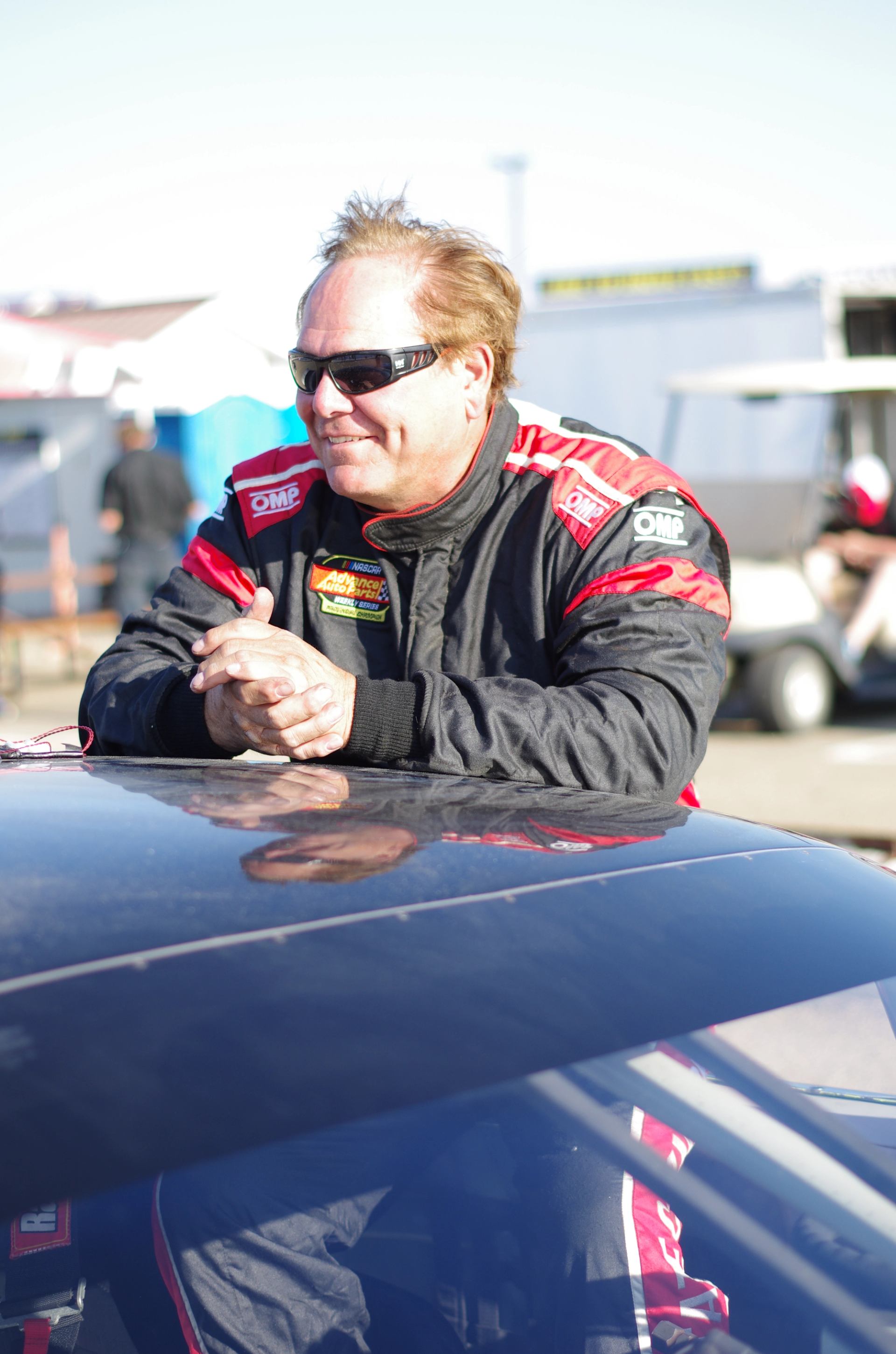 Person in a racing suit leaning on a NASCAR stock car at the track.