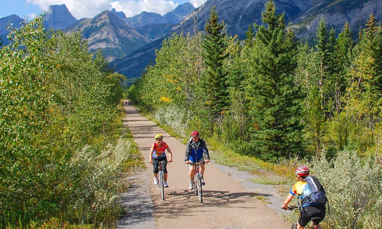 Cyclists ride along a paved forest trail with tall mountains in the background.