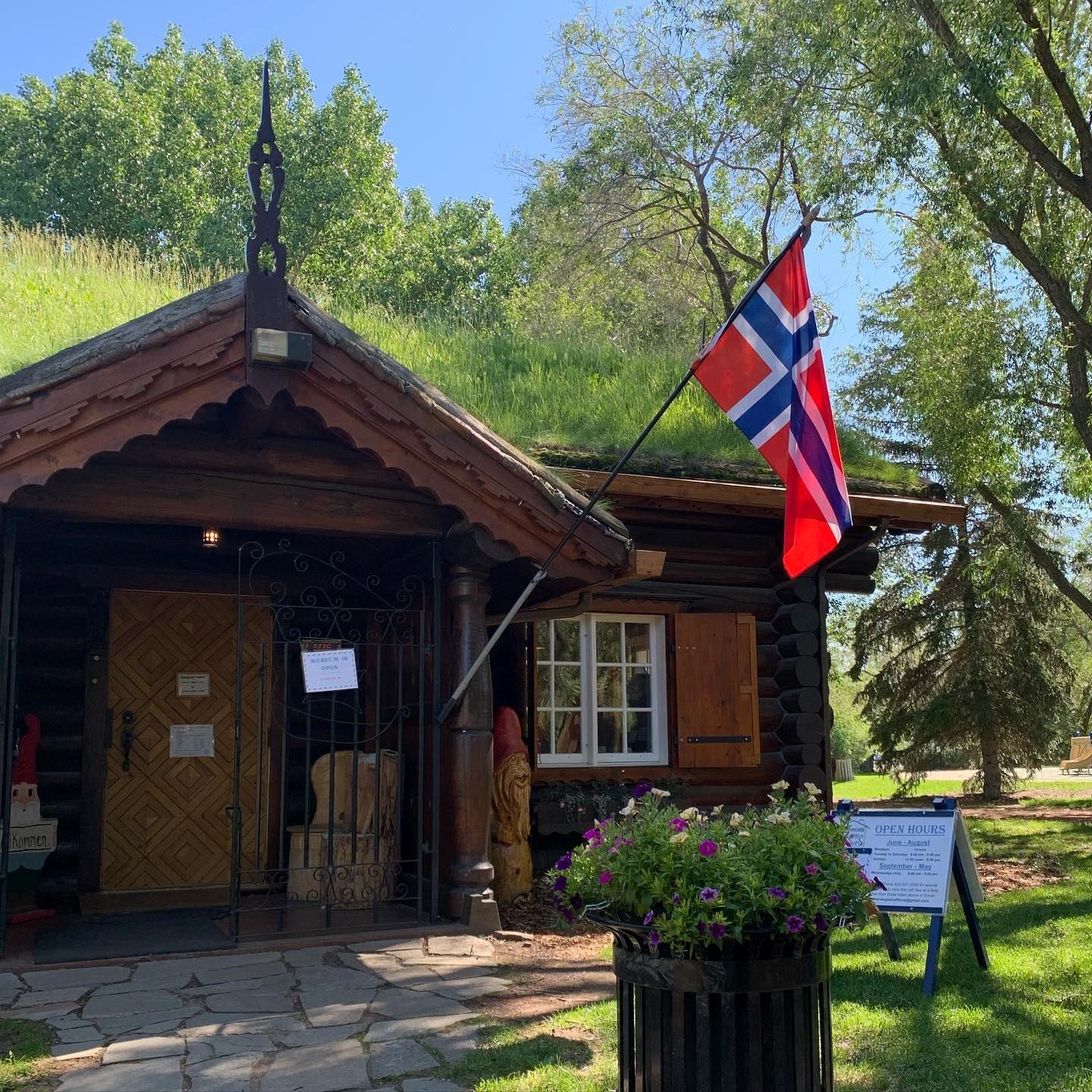 Wooden building with grass roof, Norwegian flag, and signs in a green, tree-lined setting.
