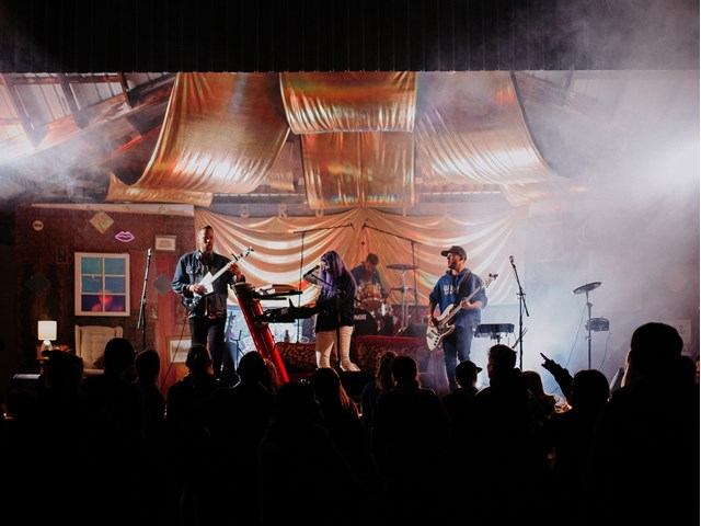 Musicians performing on stage with guitars and drums as audience silhouettes fill the foreground.