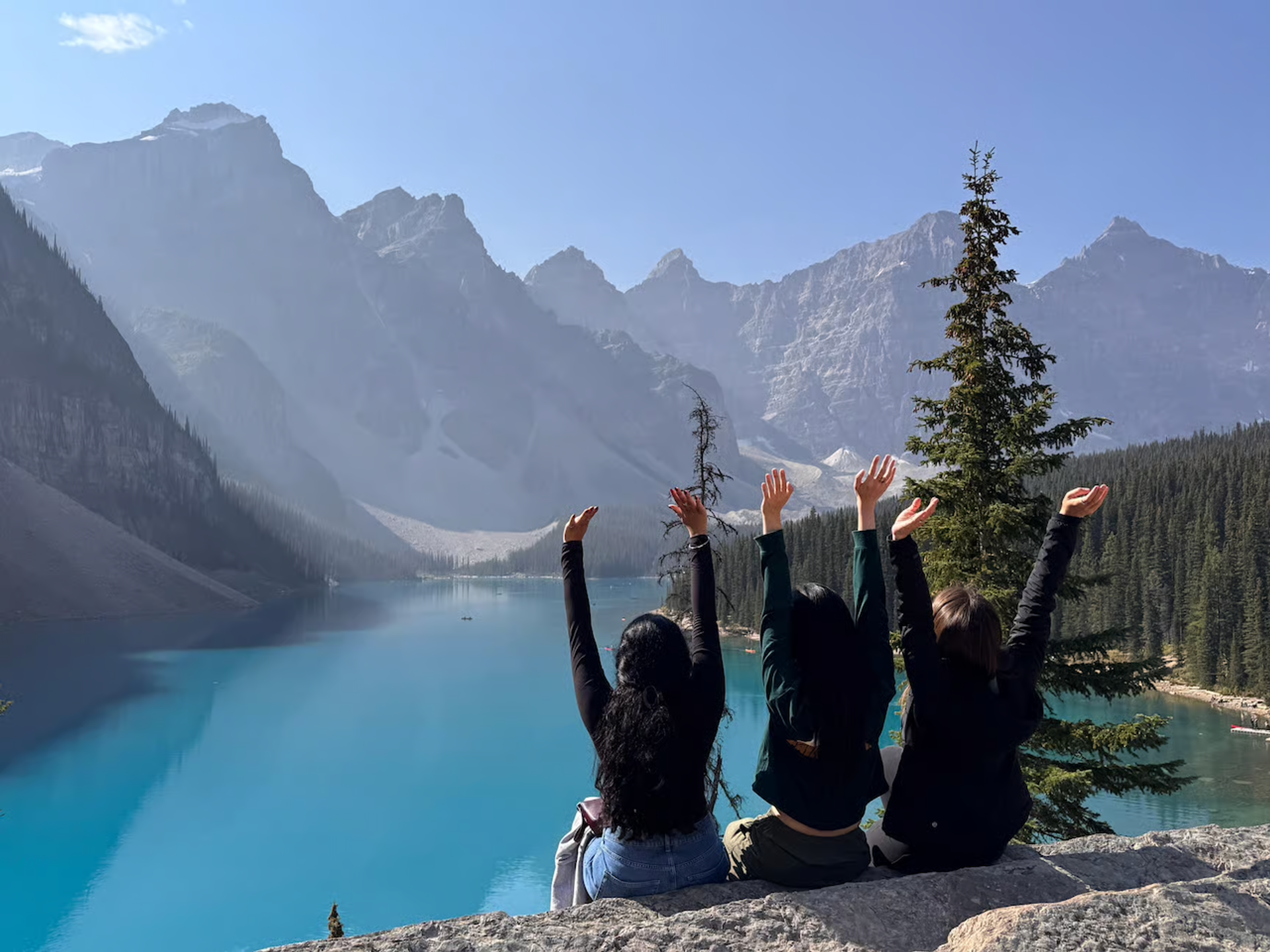 Three people sitting on a rock overlooking bright blue Moraine Lake and mountains.