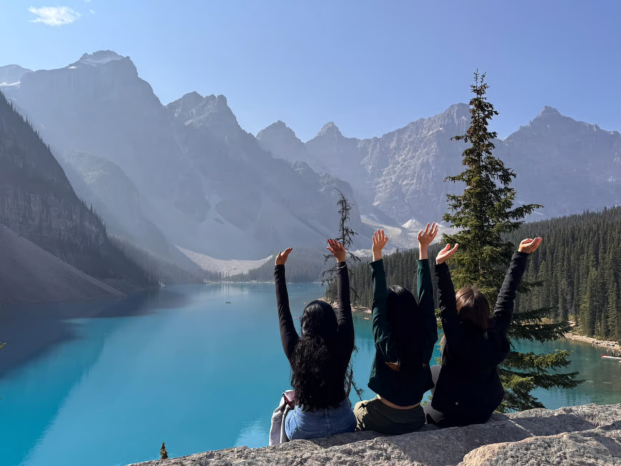 Three people sitting on a rock overlooking bright blue Moraine Lake and mountains.