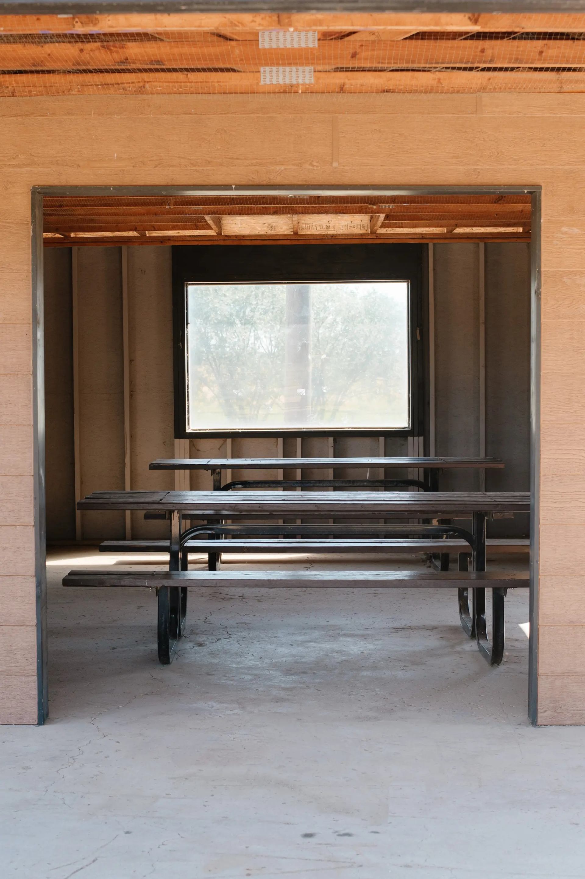 Empty wooden picnic tables inside unfinished shelter with exposed framing and large window.